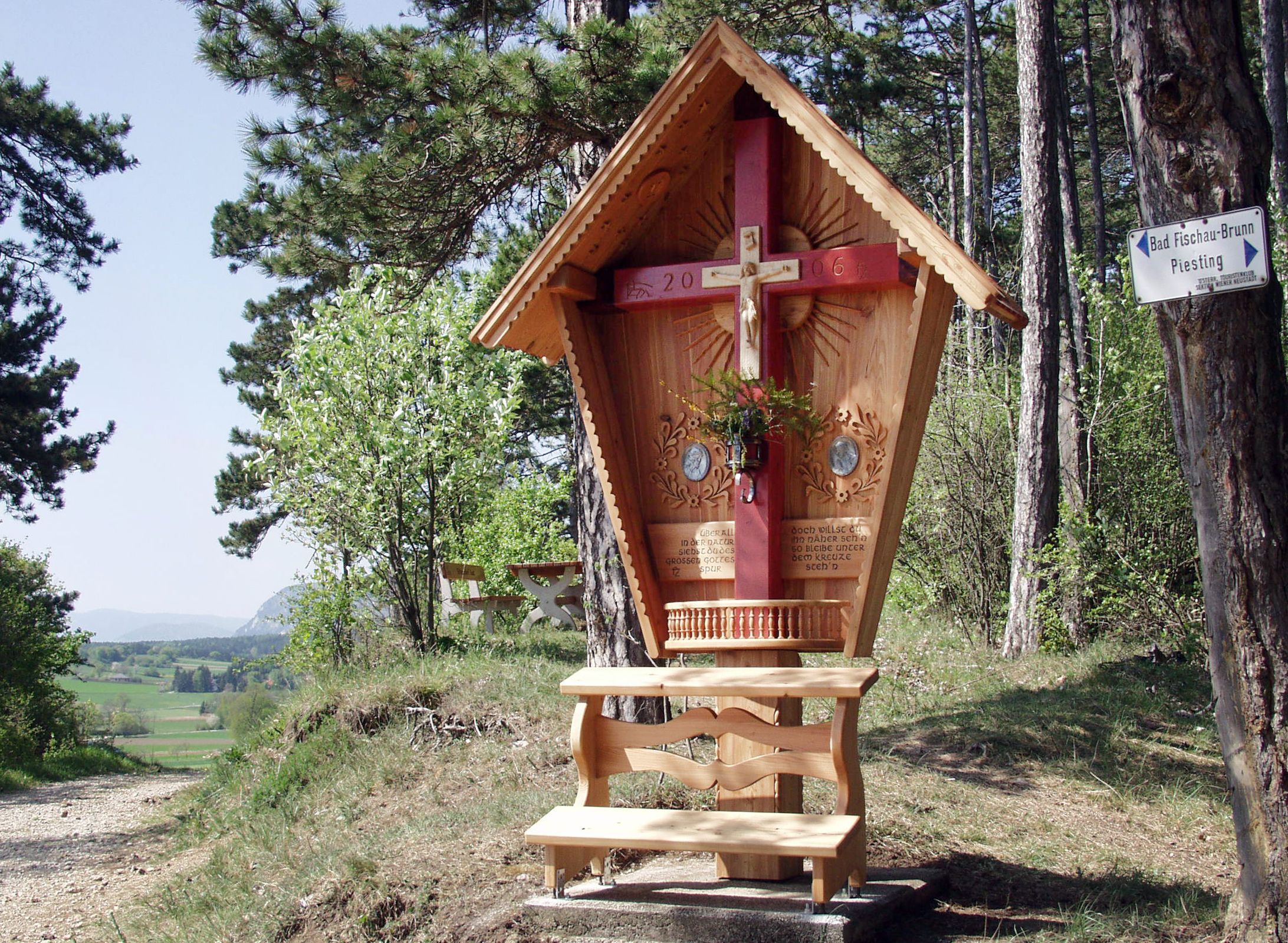 Dřevěný kříž s červeným trámem a vyřezávanou střechou v lesní krajině.