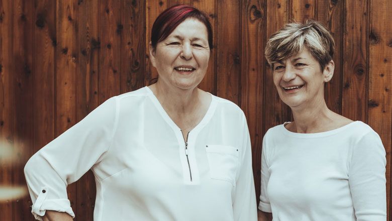 Dvě ženy v bílých halenkách stojí před dřevěnou stěnou a usmívají se.