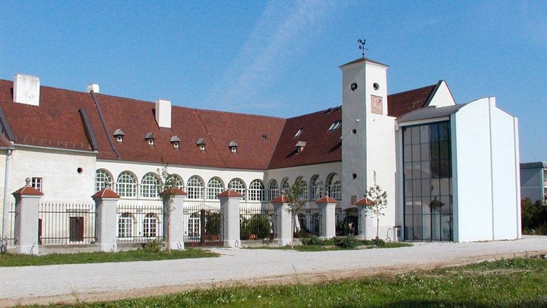 Zámek Katzelsdorf s moderní přístavbou a historickou budovou. Bílá fasáda se zaoblenými okny, červená střecha a velké vnitřní nádvoří.