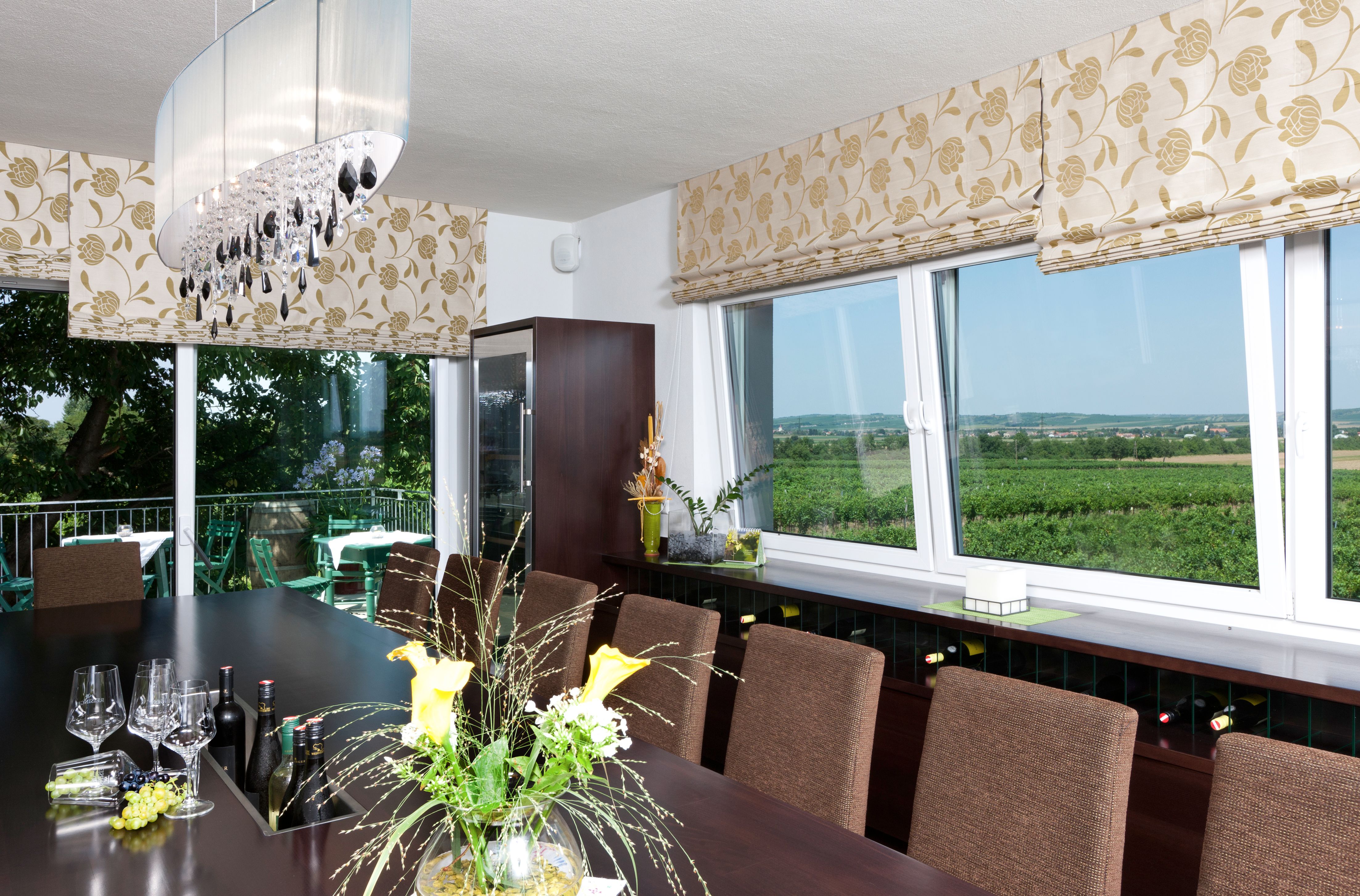 Degustační místnost s velkým stolem, sklenicemi a lahvemi vína. Velká okna nabízejí výhled na vinice. Elegantní výzdoba a osvětlení.