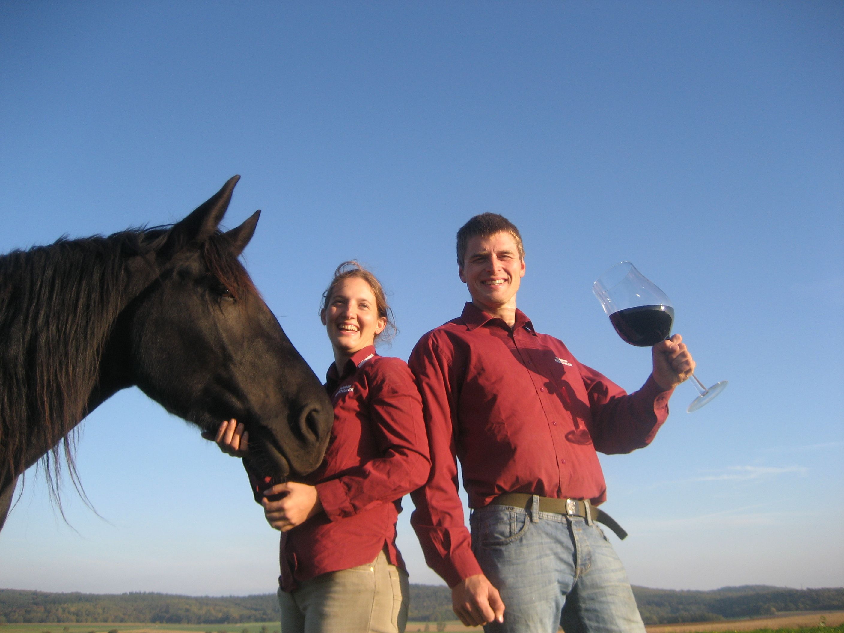 Dvě osoby v červených košilích s černým koněm a velkou sklenicí vína venku.