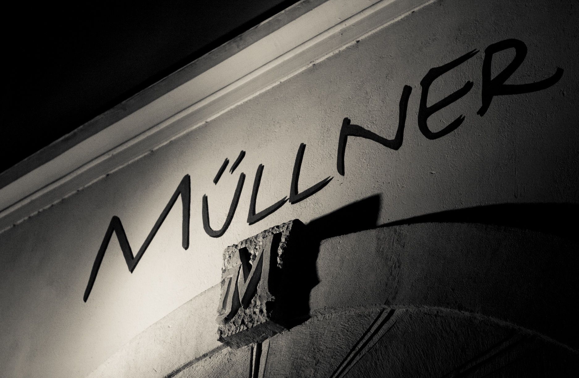 Černobílá fotografie nápisu "Müllner" na zdi s ozdobným písmenem "M".