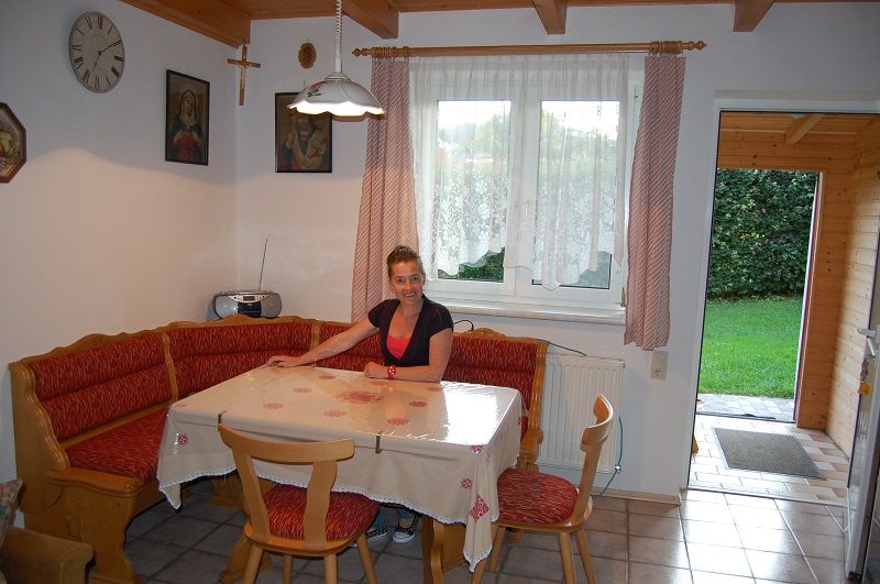 Žena sedí u jídelního stolu v útulné kuchyni s rohovou lavicí a červenými polštáři.
