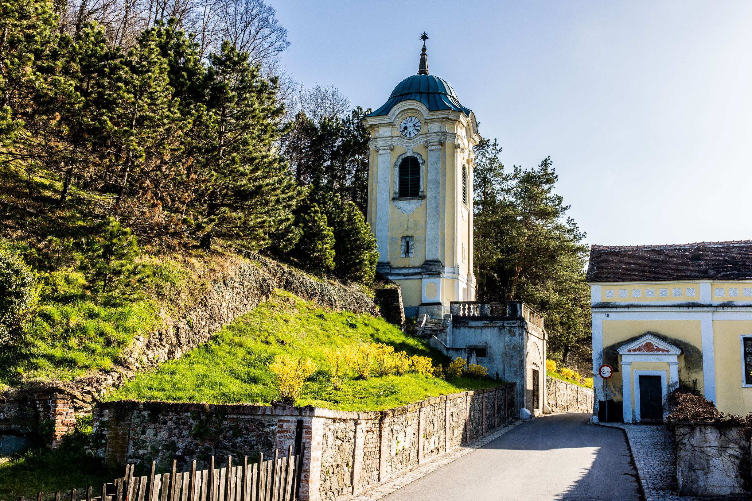 Žlutá věž s hodinami v Bad Erlachu, obklopená stromy a slunnou strání, kolem které vede úzká silnice.