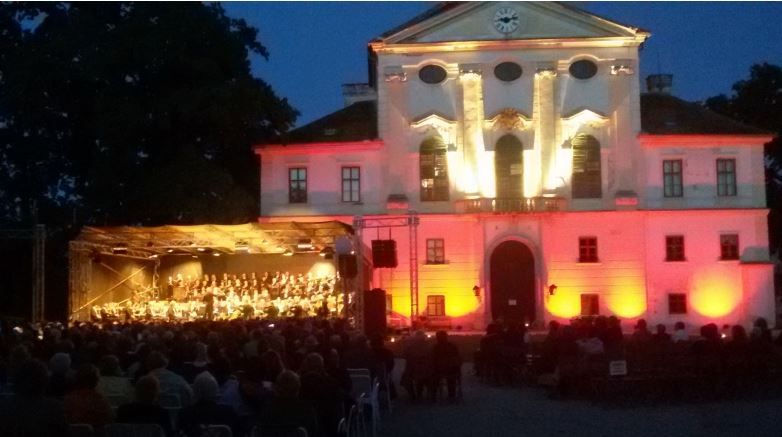 Osvětlená budova s orchestrem hrajícím na venkovním pódiu, obklopená sedícím publikem v noci.