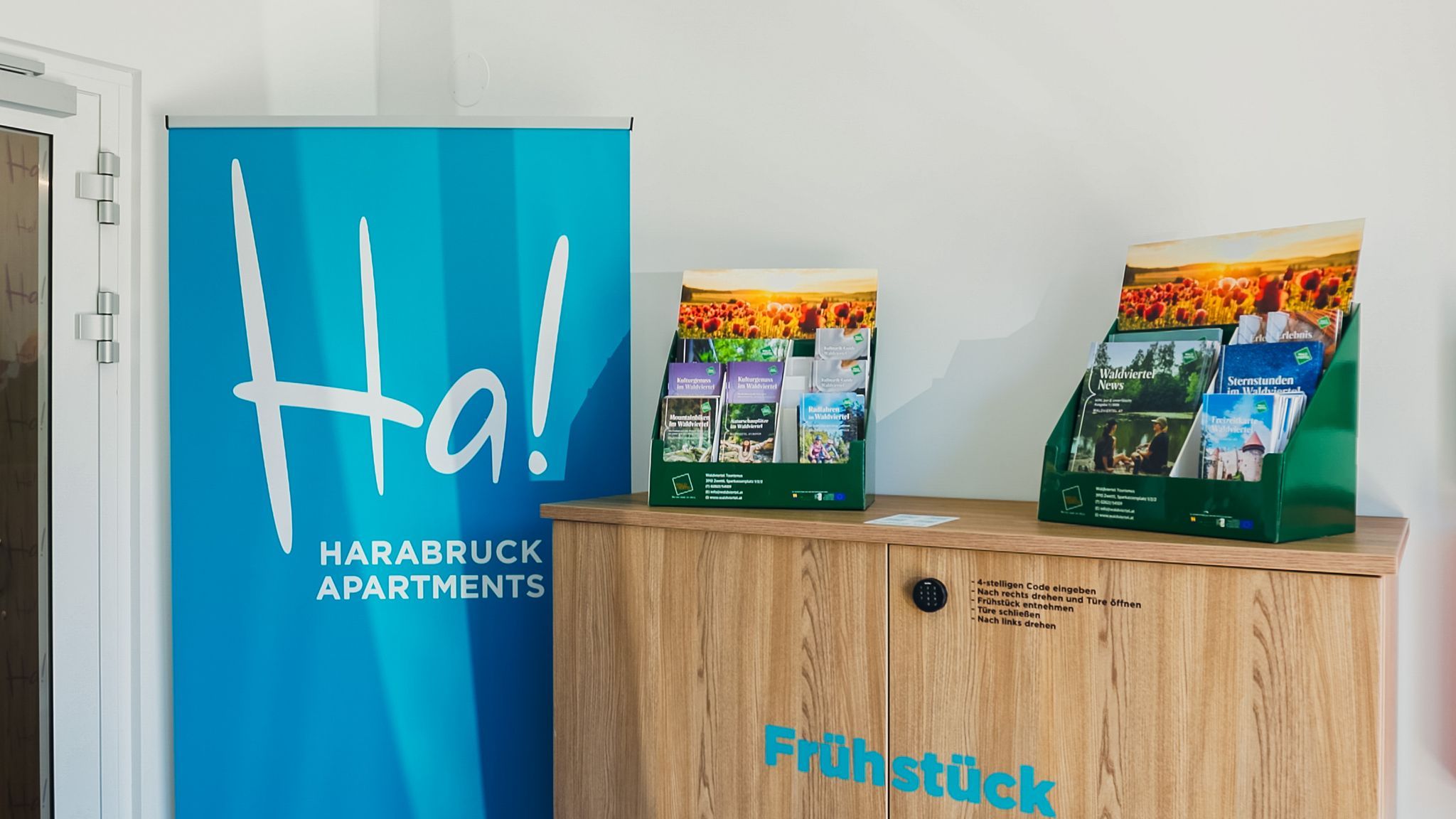 Recepční prostor s modrým bannerem Harabruck Apartments a stojanem na prospekty umístěným na dřevěné skříni.