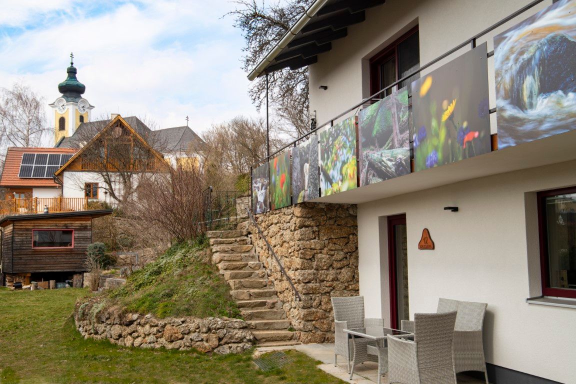Moderní dům s balkonem a výhledem do přírody, vedle něj kostel s cibulovitou věží a další budova se solárními panely.