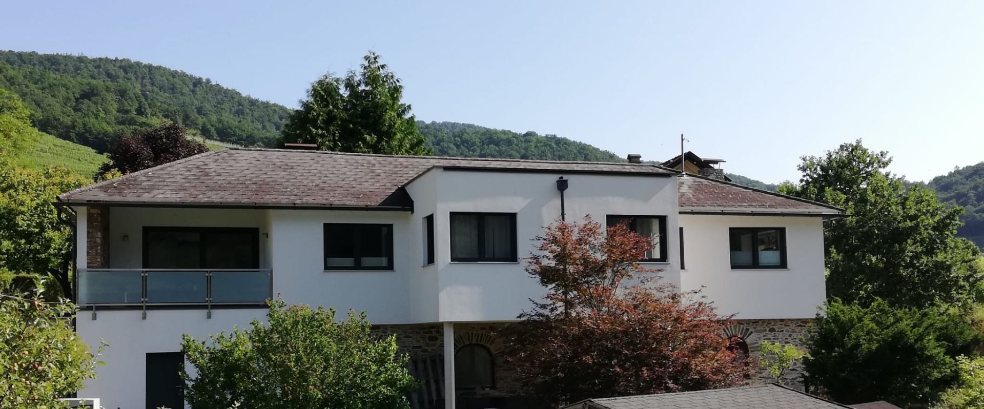 Pohled zvenčí na moderní bílý rekreační dům se stromy v popředí a zalesněnými kopci v pozadí.