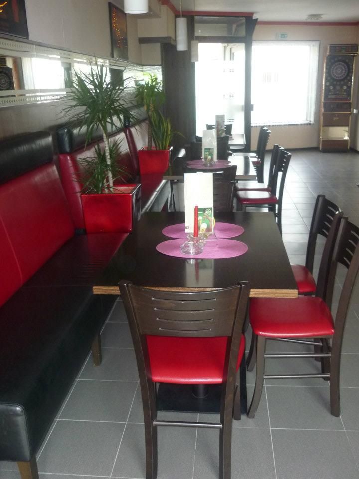 Pohled do interiéru kavárny s červeným a černým nábytkem, rostlinami a automatem na šipky.