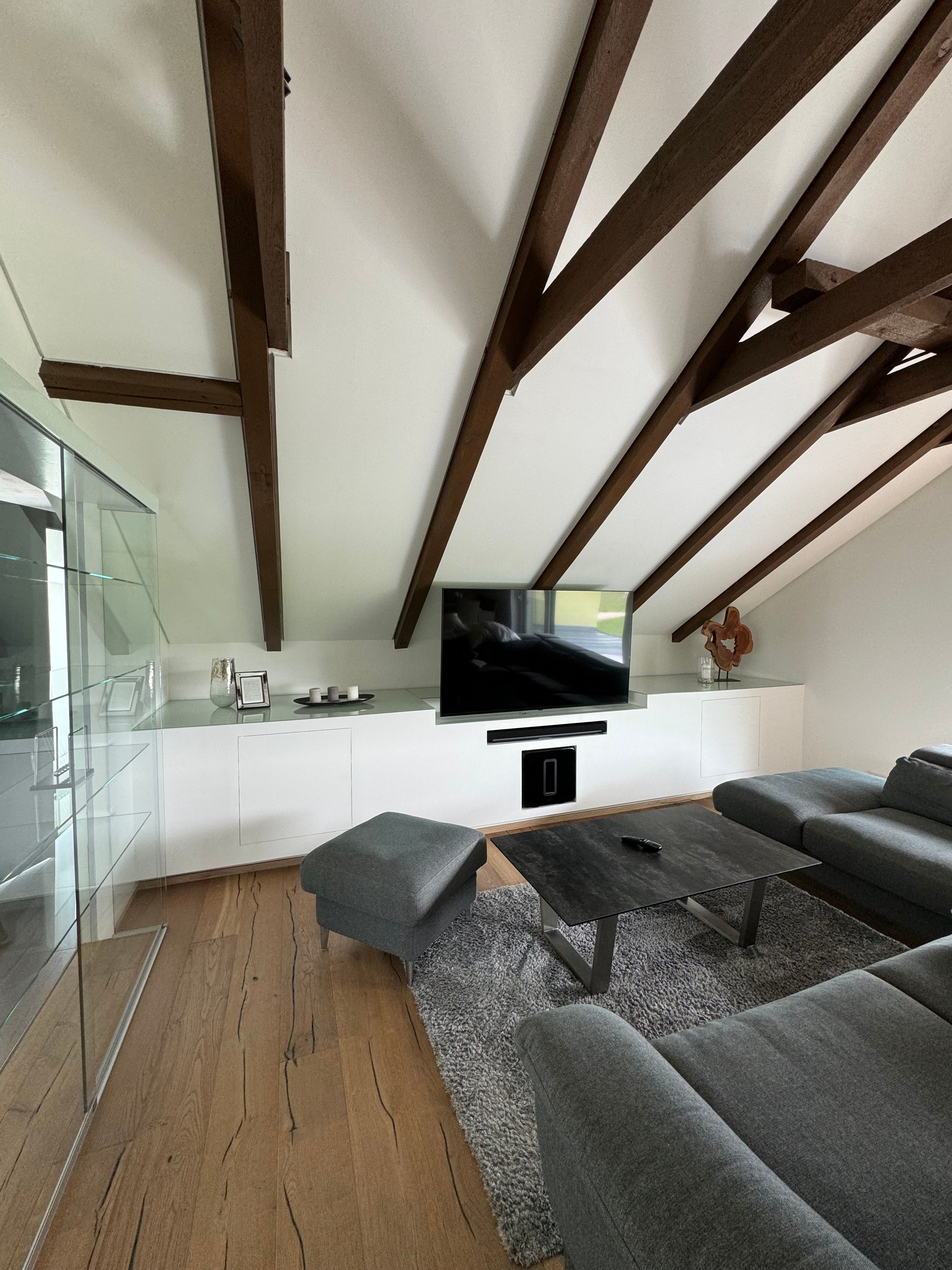 Moderní obývací prostor s šedou pohovkou, dřevěným trámovým stropem a TV s plochou obrazovkou.