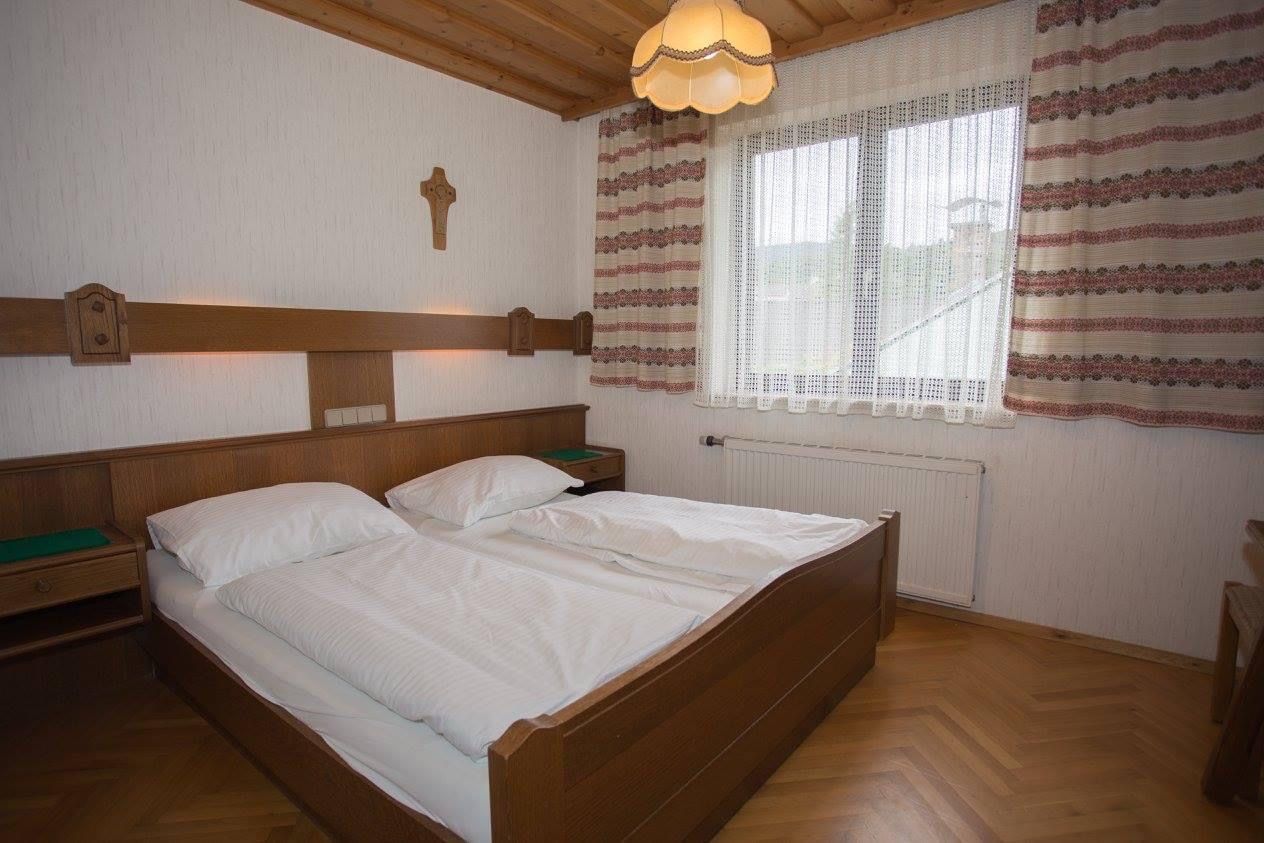 Tradičně zařízený dvoulůžkový pokoj s dřevěným nábytkem, manželskou postelí, křížem na zdi a oknem se závěsy.