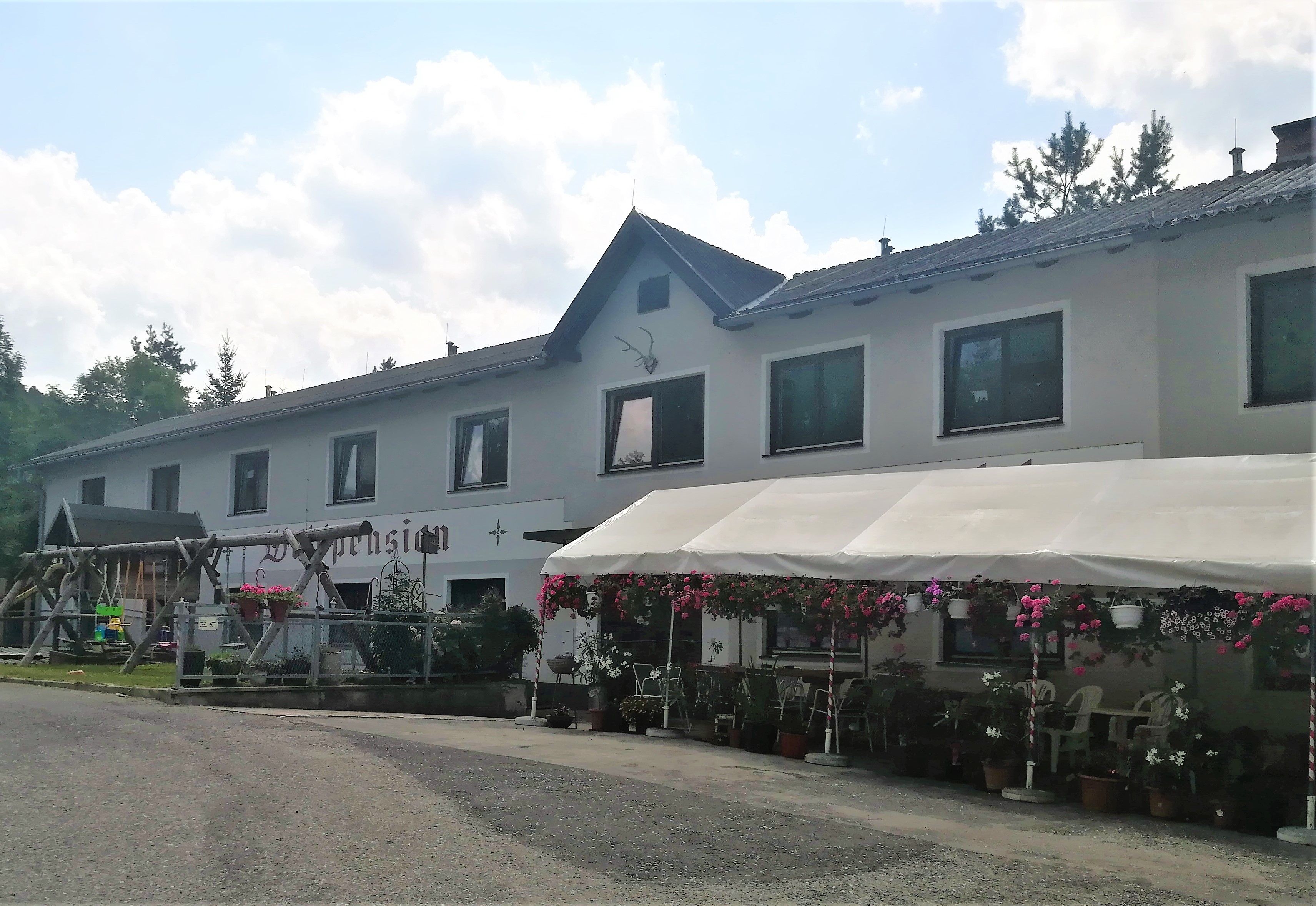 Dvoupatrová budova s nápisem "Waldpension", obklopená květinami a stříškou.