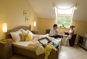 Útulný hotelový pokoj s párem sedícím u stolu. Postel je rozestlaná a na posteli leží otevřený kufr.