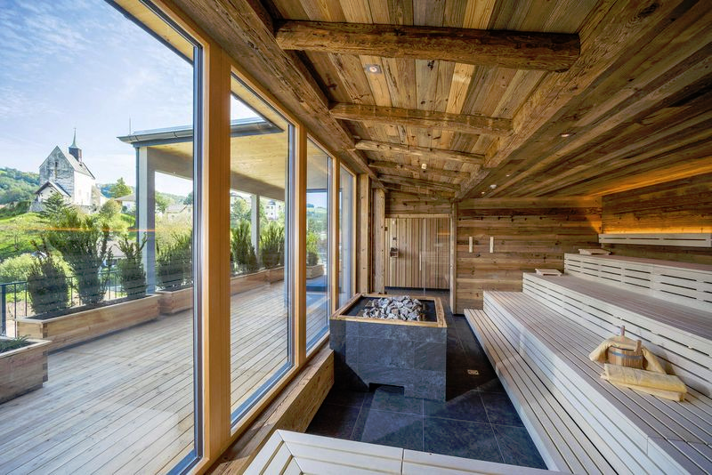 Moderní sauna s dřevěným obložením, tmavými dlaždicemi a proskleným průčelím s výhledem ven.