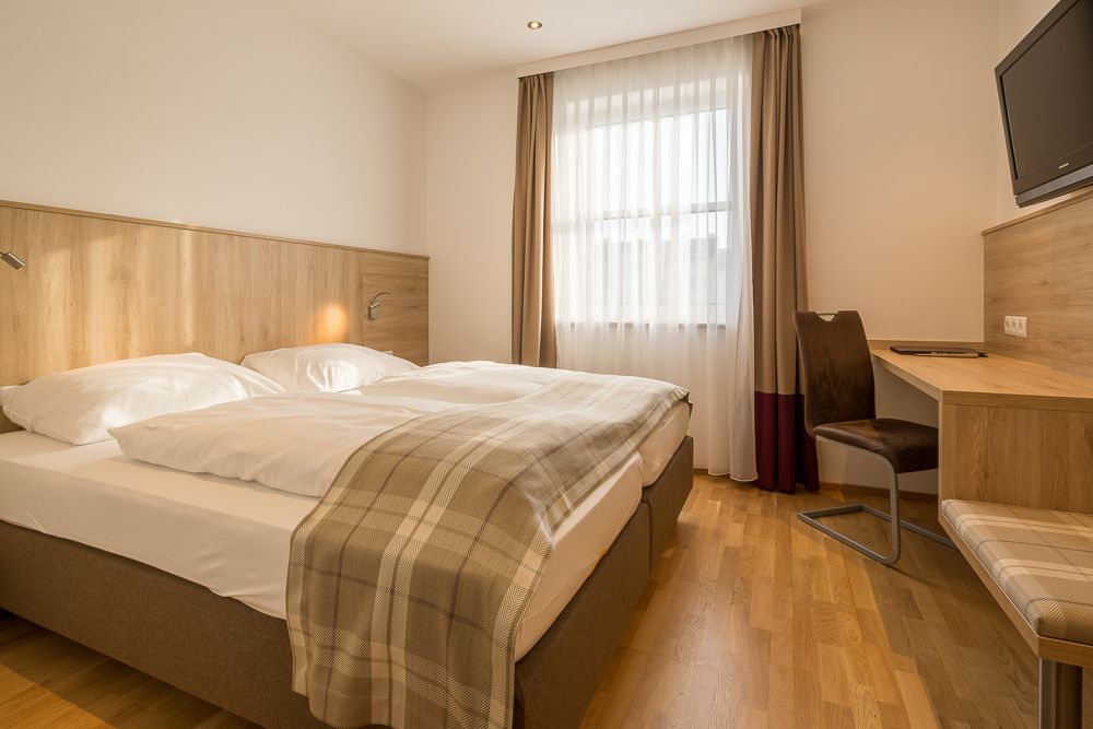 Hotelový pokoj s manželskou postelí, psacím stolem a oknem se závěsy.