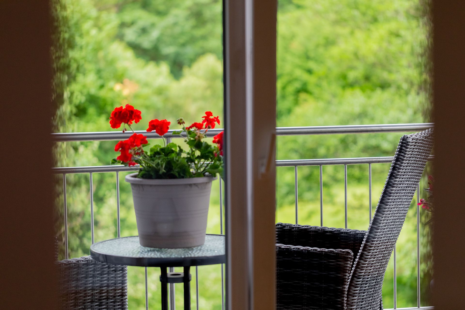 Pohled na balkon se stolem, květináčem s červenými květy a židlí, obklopený zelení.