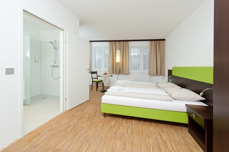 Moderní hotelový pokoj s manželskou postelí, dřevěnou podlahou a přilehlou koupelnou.