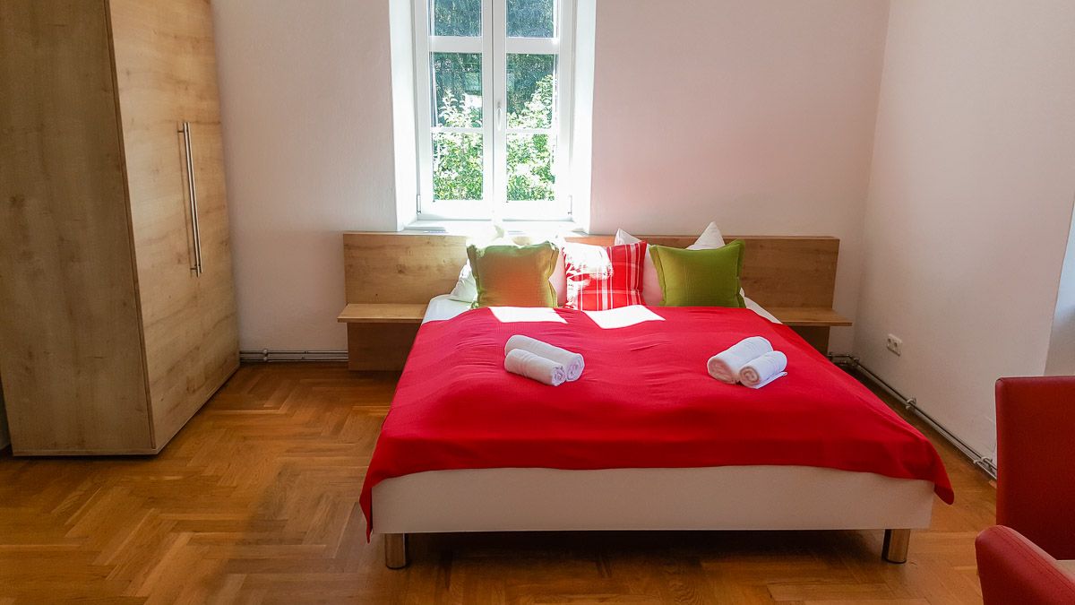 Ložnice s červeným přehozem, polštáři a ručníky na posteli, dřevěnou skříní a oknem v pozadí.