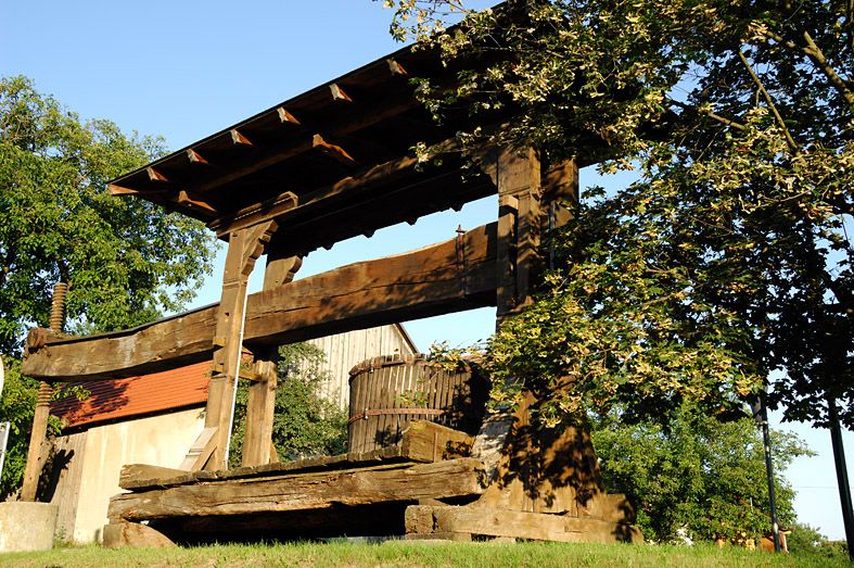 Starý dřevěný lis na víno pod širým nebem, obklopený stromy a budovami.