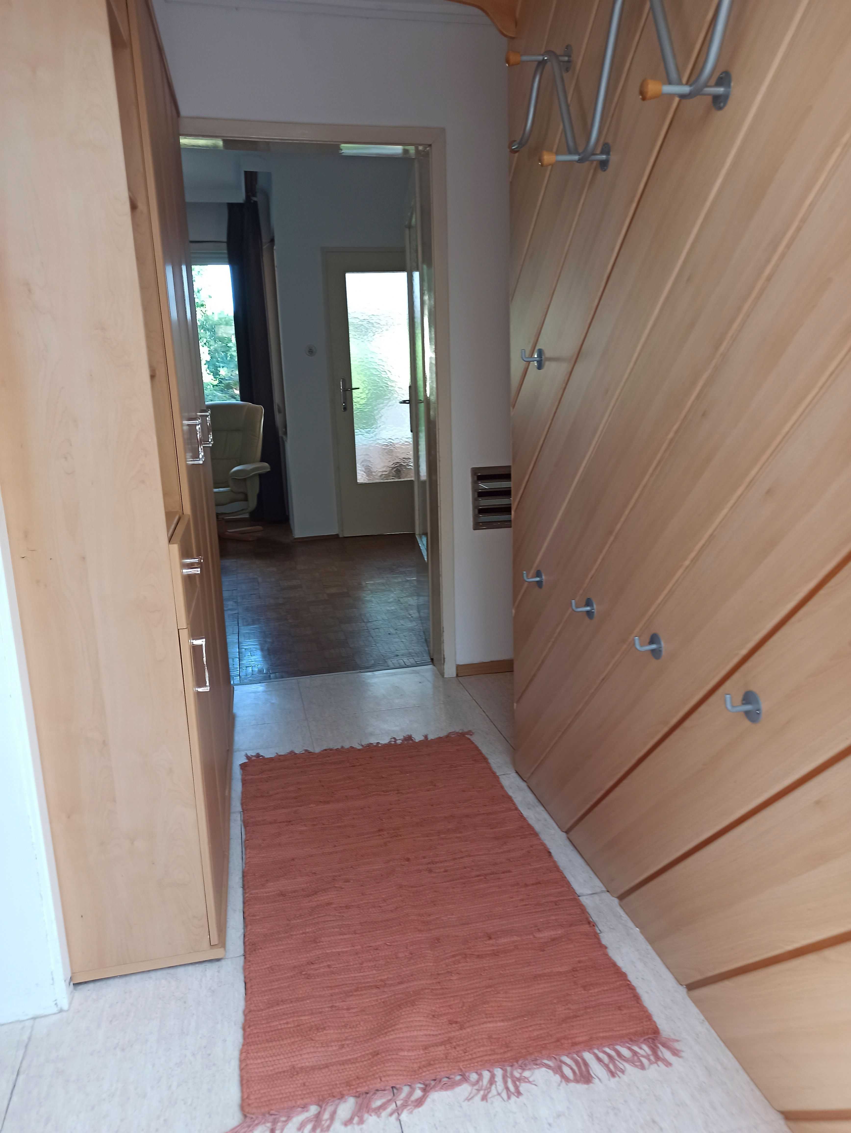 Chodba s dřevěnými stěnami, háčky a červeným kobercem. V pozadí je vidět pokoj s křeslem a skleněnými dveřmi.