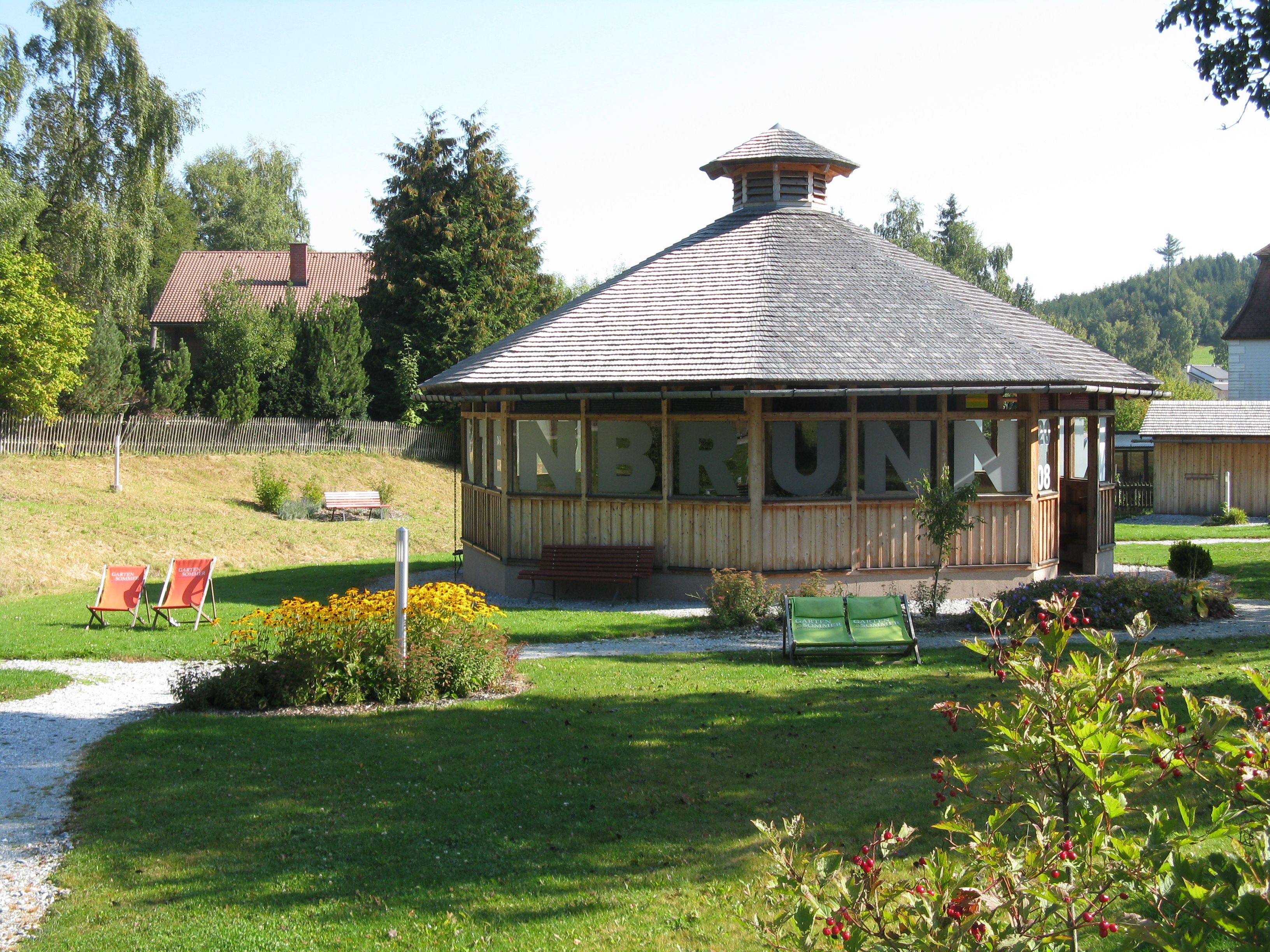 Pavilon v zeleném parku s květinami a lehátky.