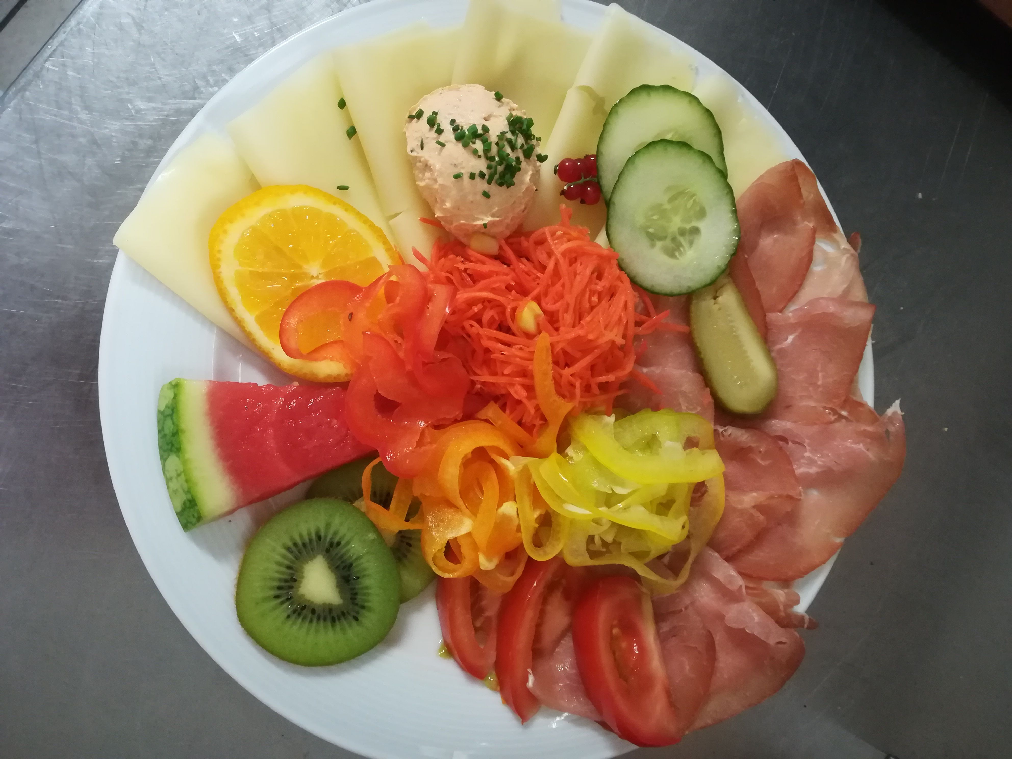 Talíř se sýrem, šunkou, zeleninou a ovocem, včetně melounu, kiwi, okurky a papriky.