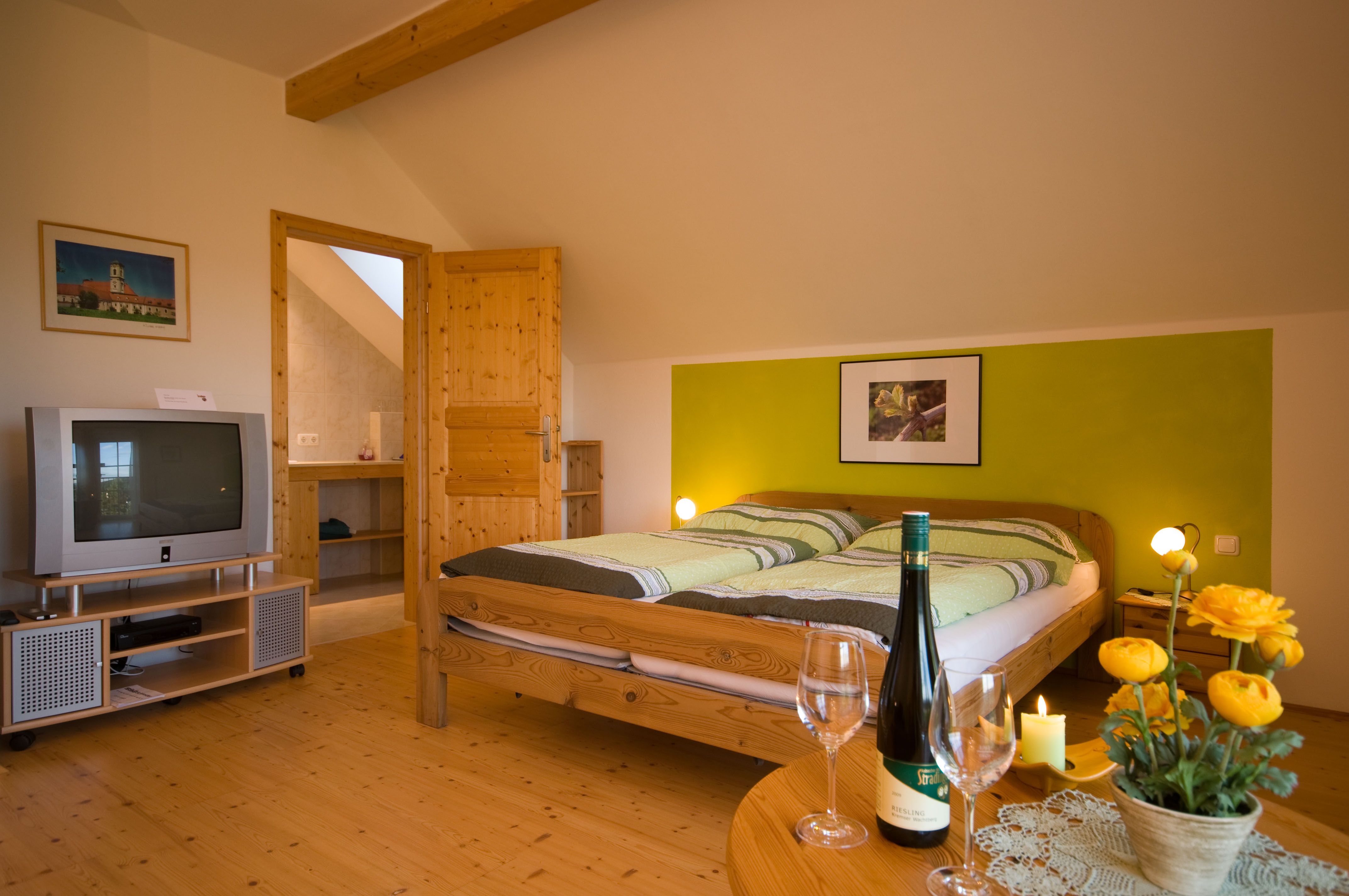 Útulný pokoj s manželskou postelí, TV, lahví vína a květinami na stole.