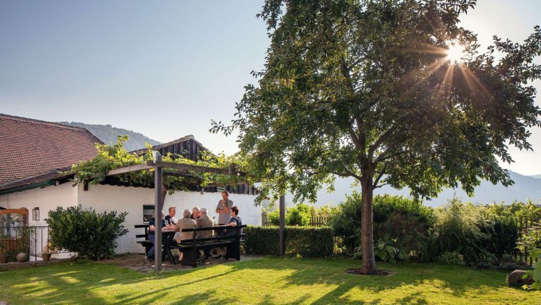 Lid&eacute; sed&iacute; u stolu na zahradě pod pergolou, obklopeni př&iacute;rodou a sluncem.