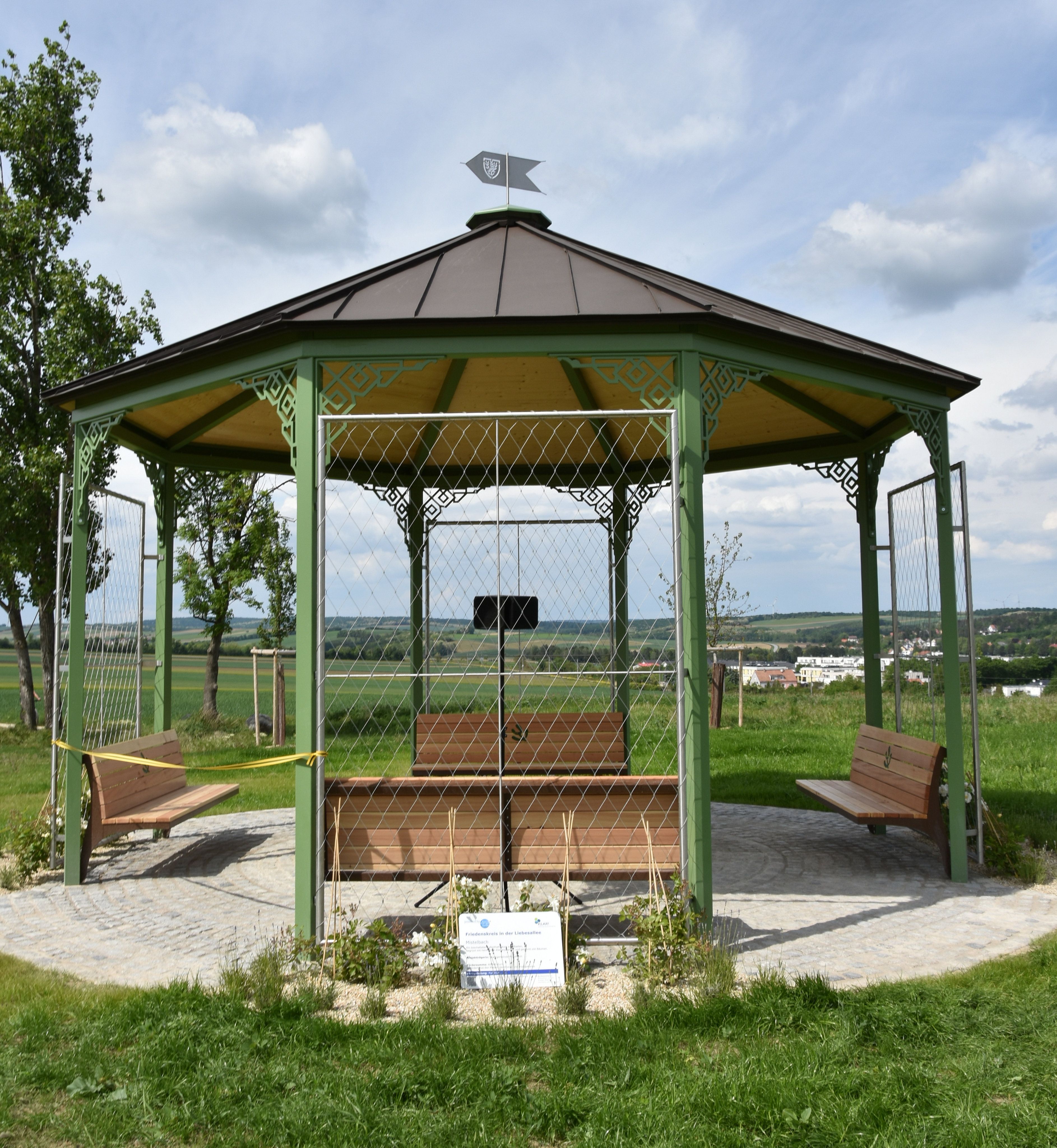 Pavilon se zelenou kovovou konstrukcí a dřevěnou střechou stojí na dlážděném náměstí ve venkovském prostředí.