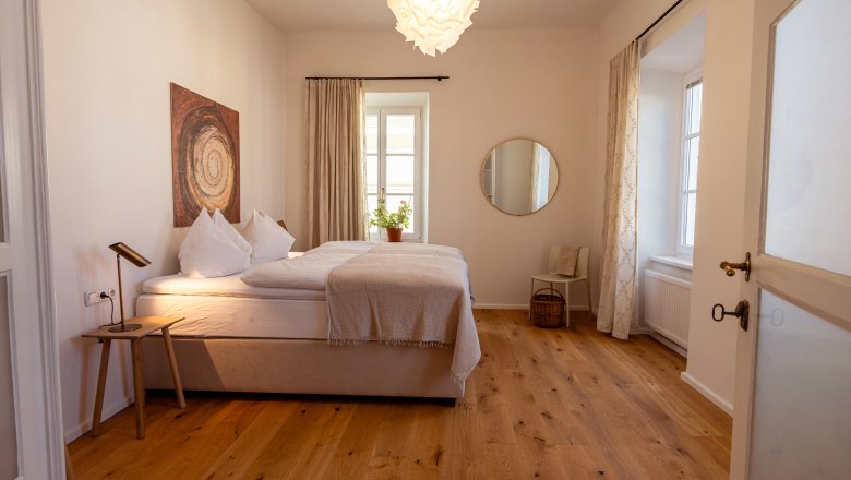 &Uacute;tuln&aacute; ložnice s manželskou postel&iacute;, dřevěnou podlahou, uměleck&yacute;mi d&iacute;ly na stěně, kulat&yacute;m zrcadlem a rostlinami na parapetu.