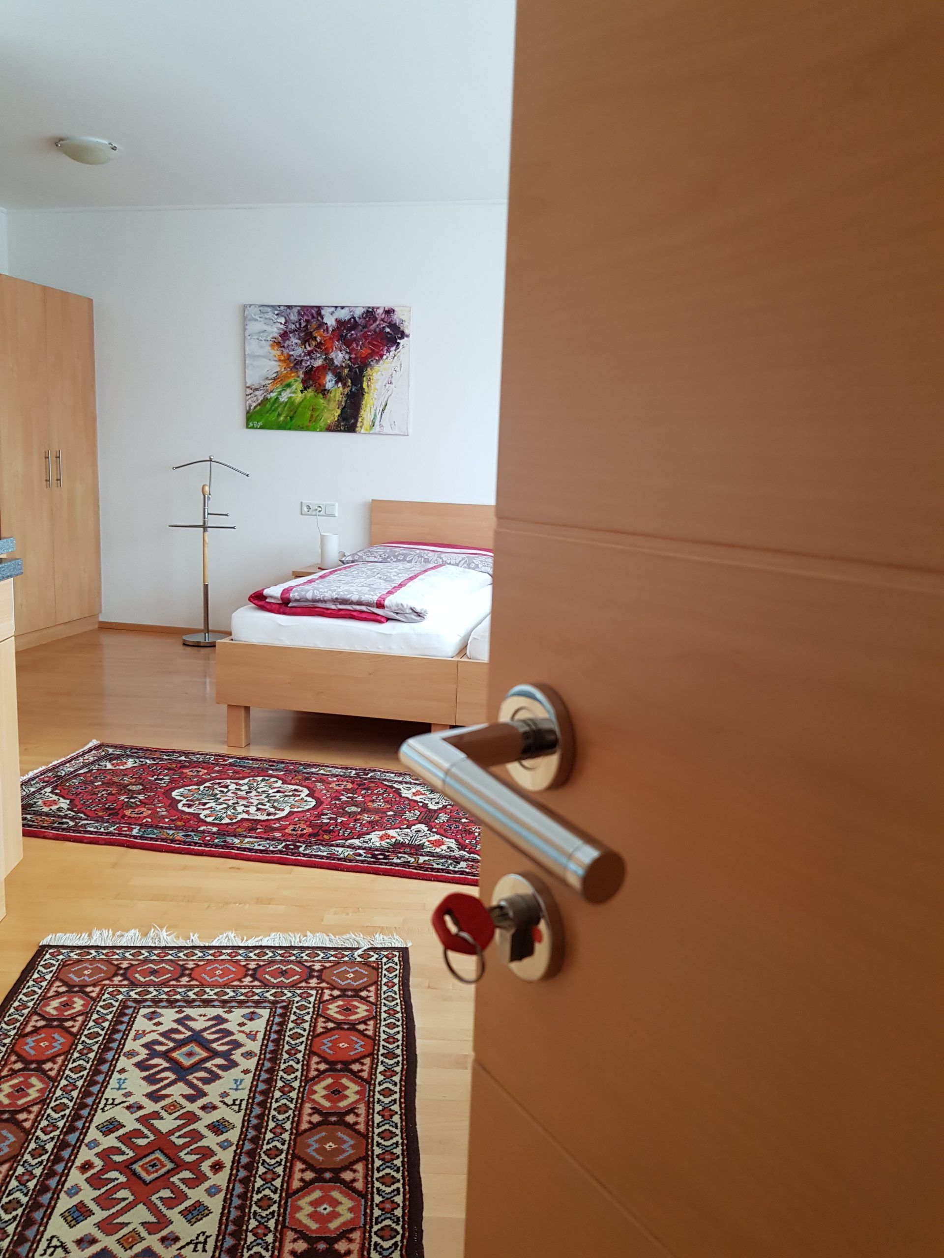 Otevřený pokoj s postelí, koberci a obrazem na stěně.