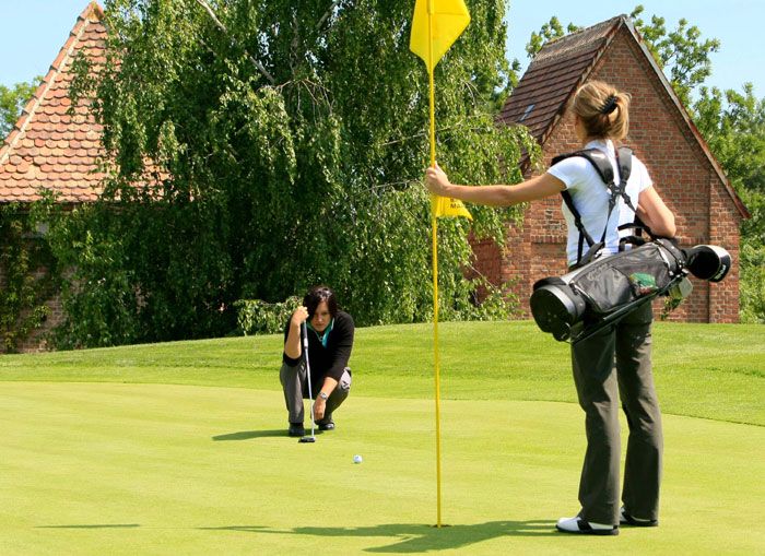 Dvě ženy hrající golf na zeleném golfovém hřišti, jedna drží vlajku, druhá míří na jamku.
