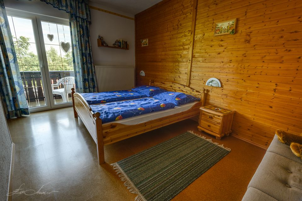 Útulná ložnice s dřevěnými stěnami, manželskou postelí, modrým povlečením a balkonem s výhledem do krajiny.