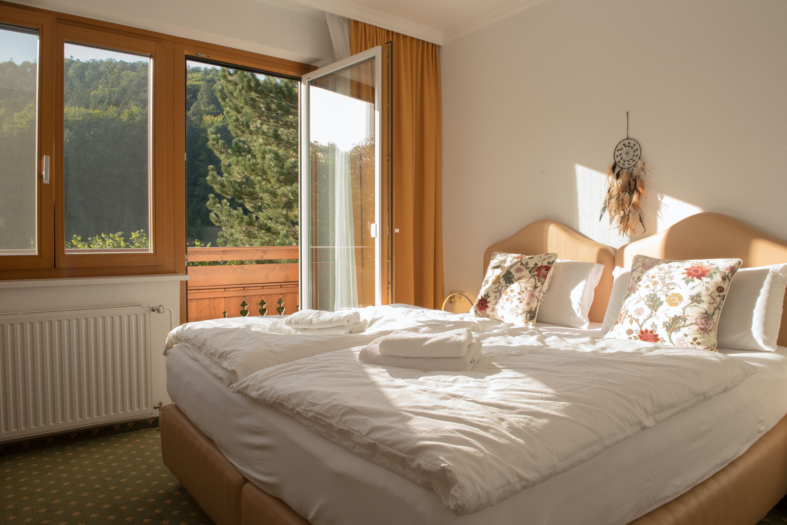 Útulný jednolůžkový pokoj s manželskou postelí, ozdobnými polštáři a otevřeným balkonem s výhledem na zelené stromy.