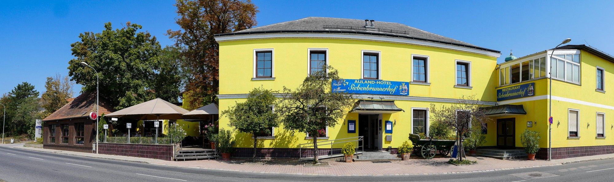 Žlutá budova s restaurací a hotelem, obklopená stromy a silnicí.