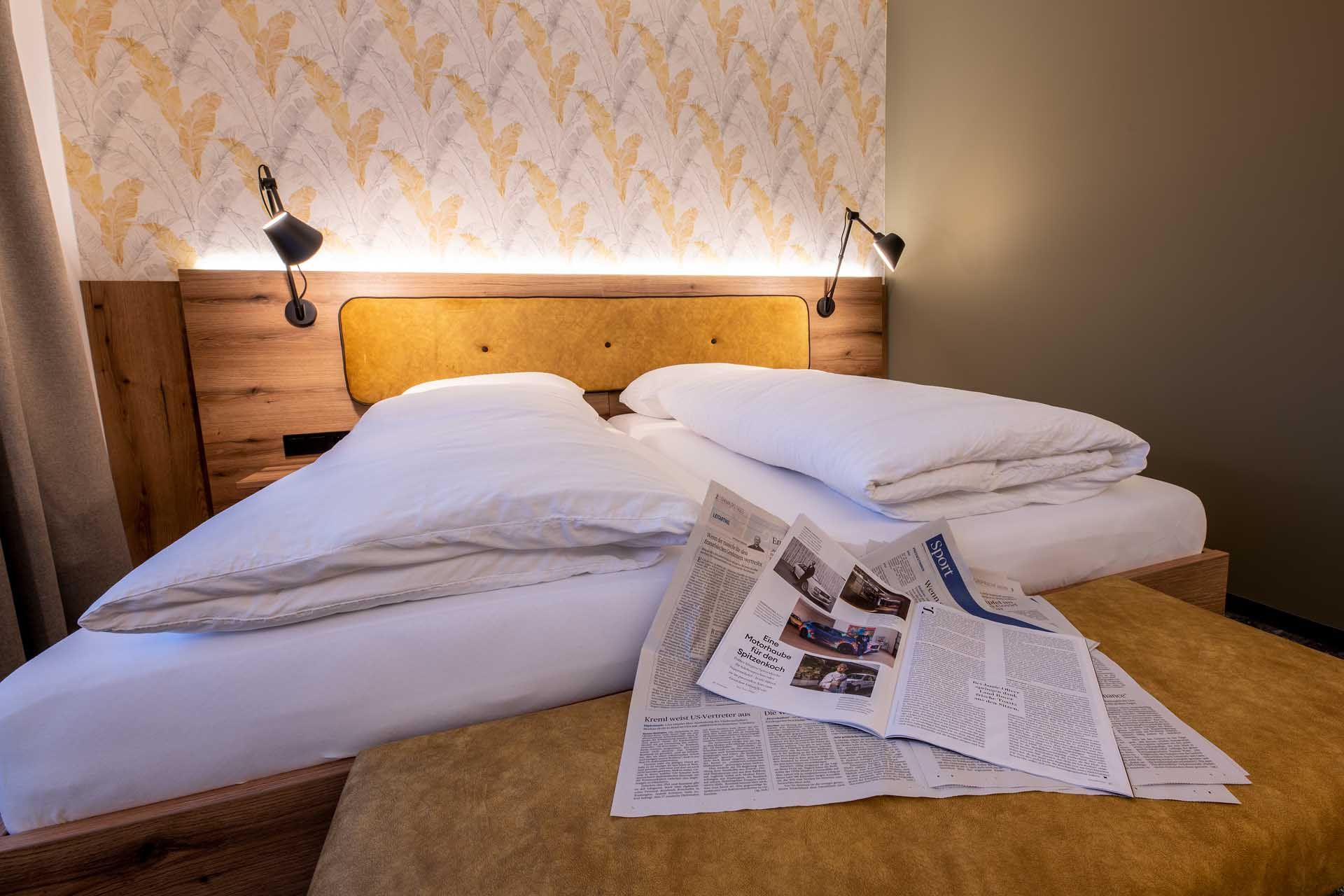 Útulný pokoj pro hosty s manželskou postelí, novinami na přikrývce a stylovou tapetou v pozadí.