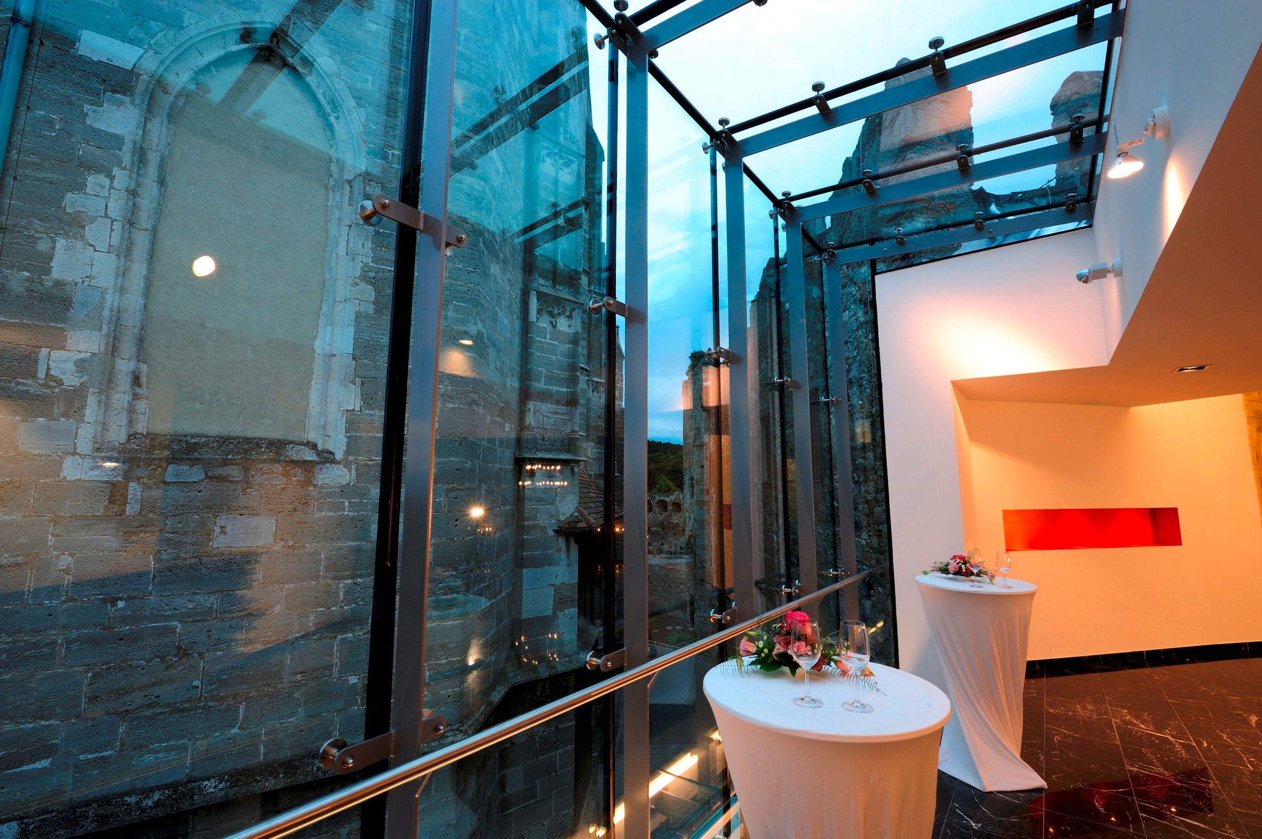 Moderní skleněná architektura vedle staré kamenné zdi se zdobenými barovými stoly.