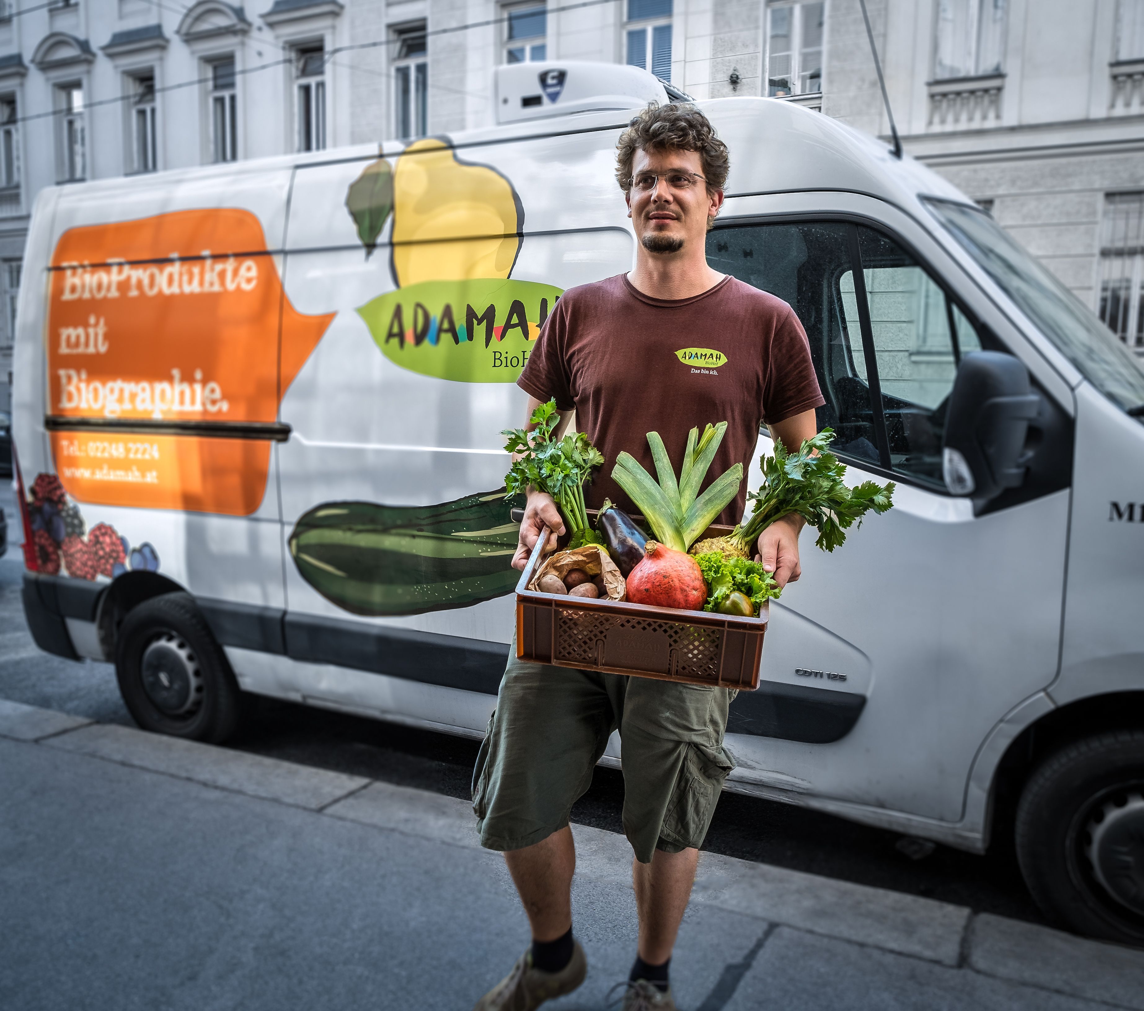 Muž drží krabici s čerstvým ovocem a zeleninou před dodávkou s logem ADAMAH.