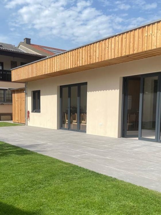 Moderní budova s dřevěným obložením a velkými okny, obklopená terasou a trávníkem.