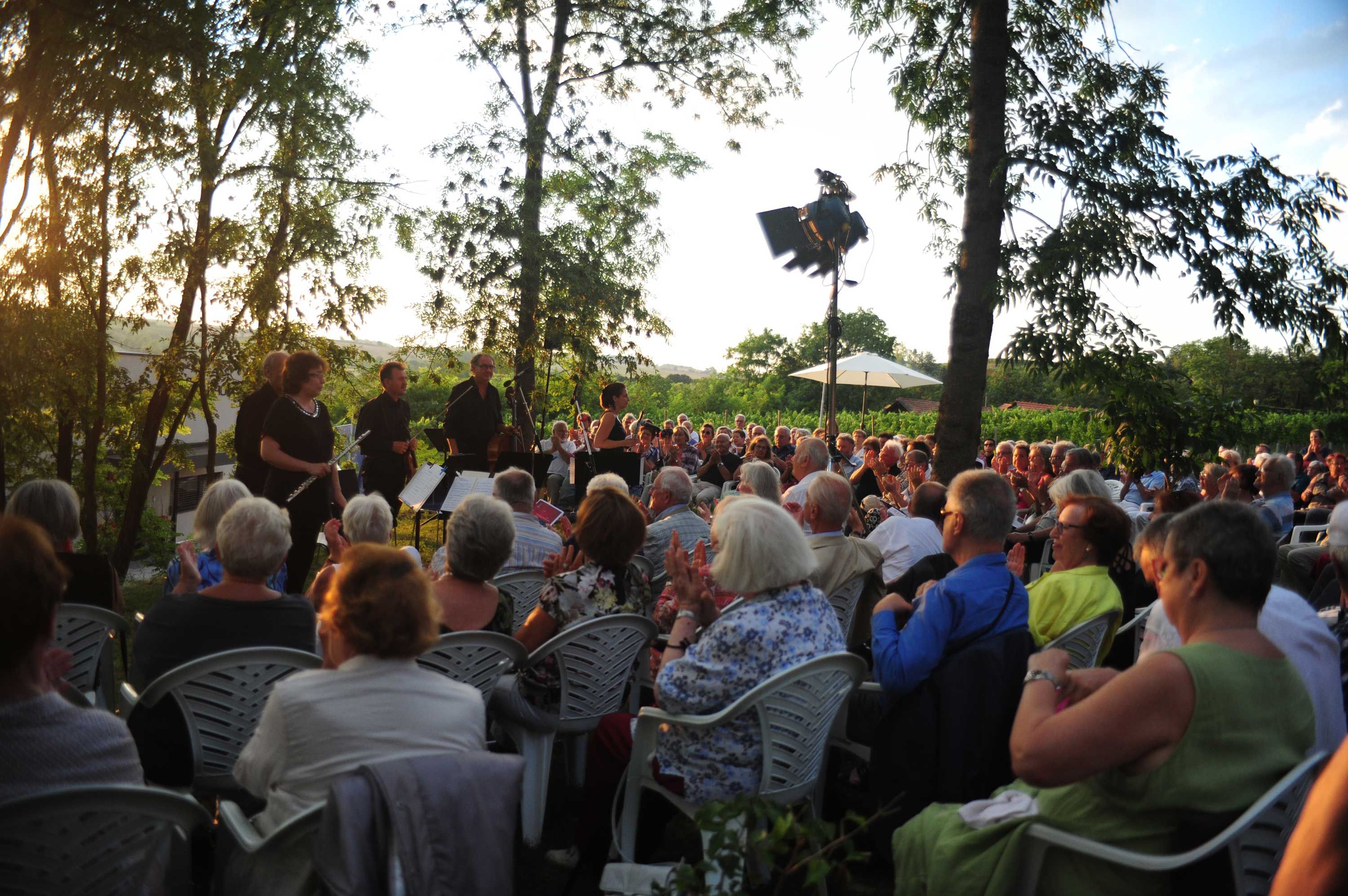 Venkovní koncert s orchestrem a publikem při západu slunce.