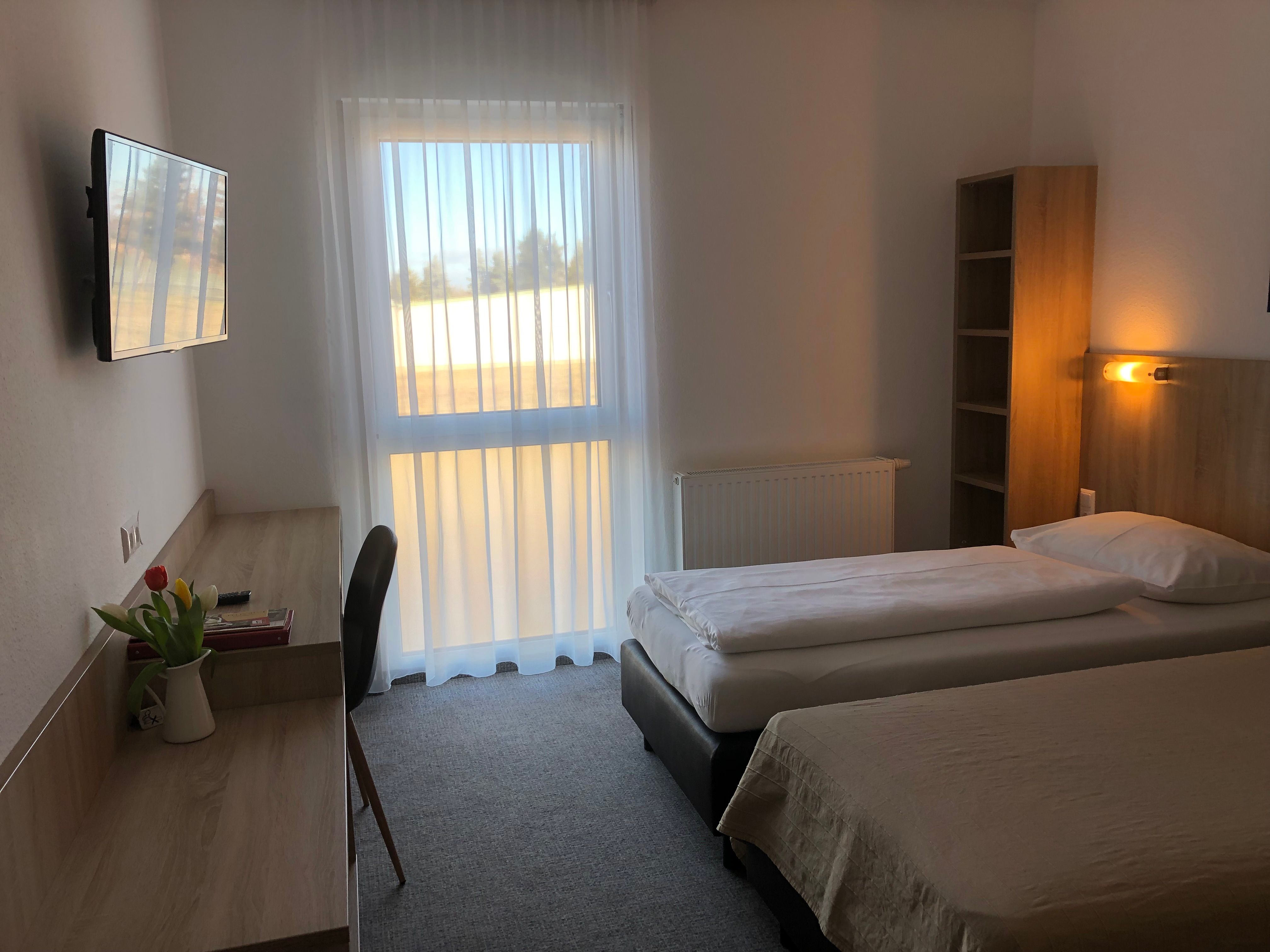 Jednoduše zařízený motelový pokoj se dvěma samostatnými lůžky, psacím stolem, televizí a oknem se závěsy.