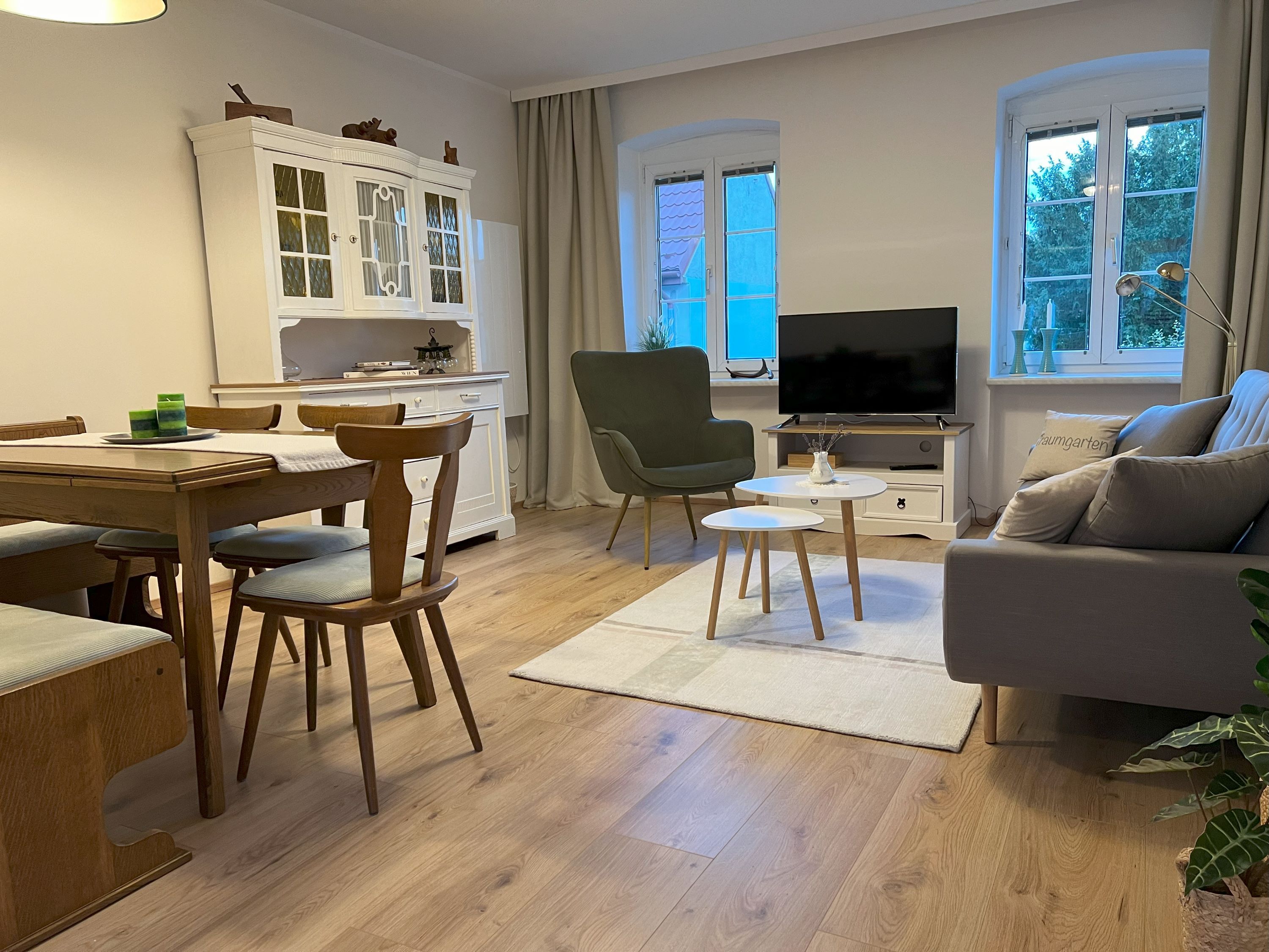 Útulný obývací pokoj s dřevěným nábytkem, pohovkou, křeslem, jídelním stolem a TV. Světlé stěny a dřevěná podlaha.