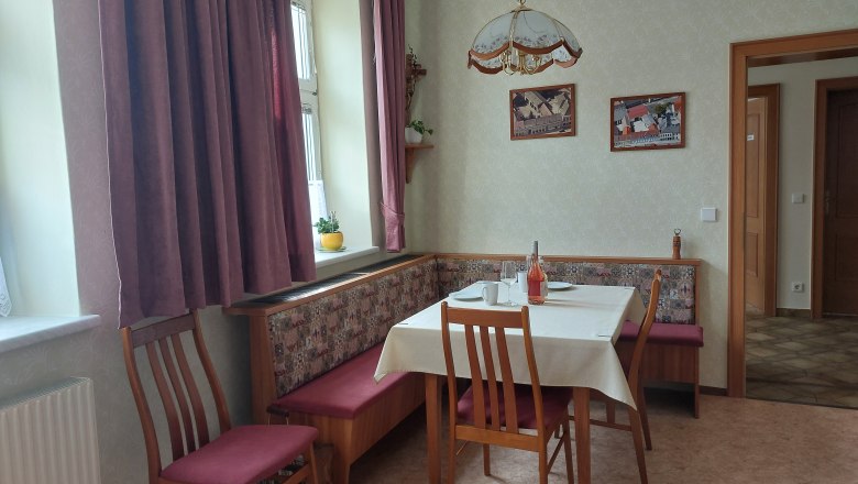 &Uacute;tuln&aacute; kuchyně s j&iacute;deln&iacute;m stolem, židlemi a rohovou lavic&iacute;. Okno se z&aacute;věsy, obr&aacute;zky na stěně.