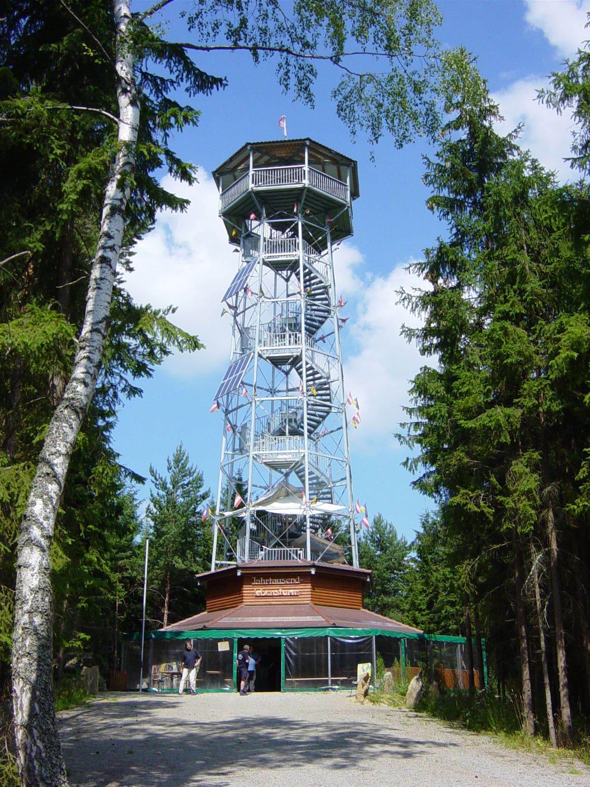 Vysoká kovová rozhledna s dřevěným obložením, obklopená stromy, pod modrou oblohou.