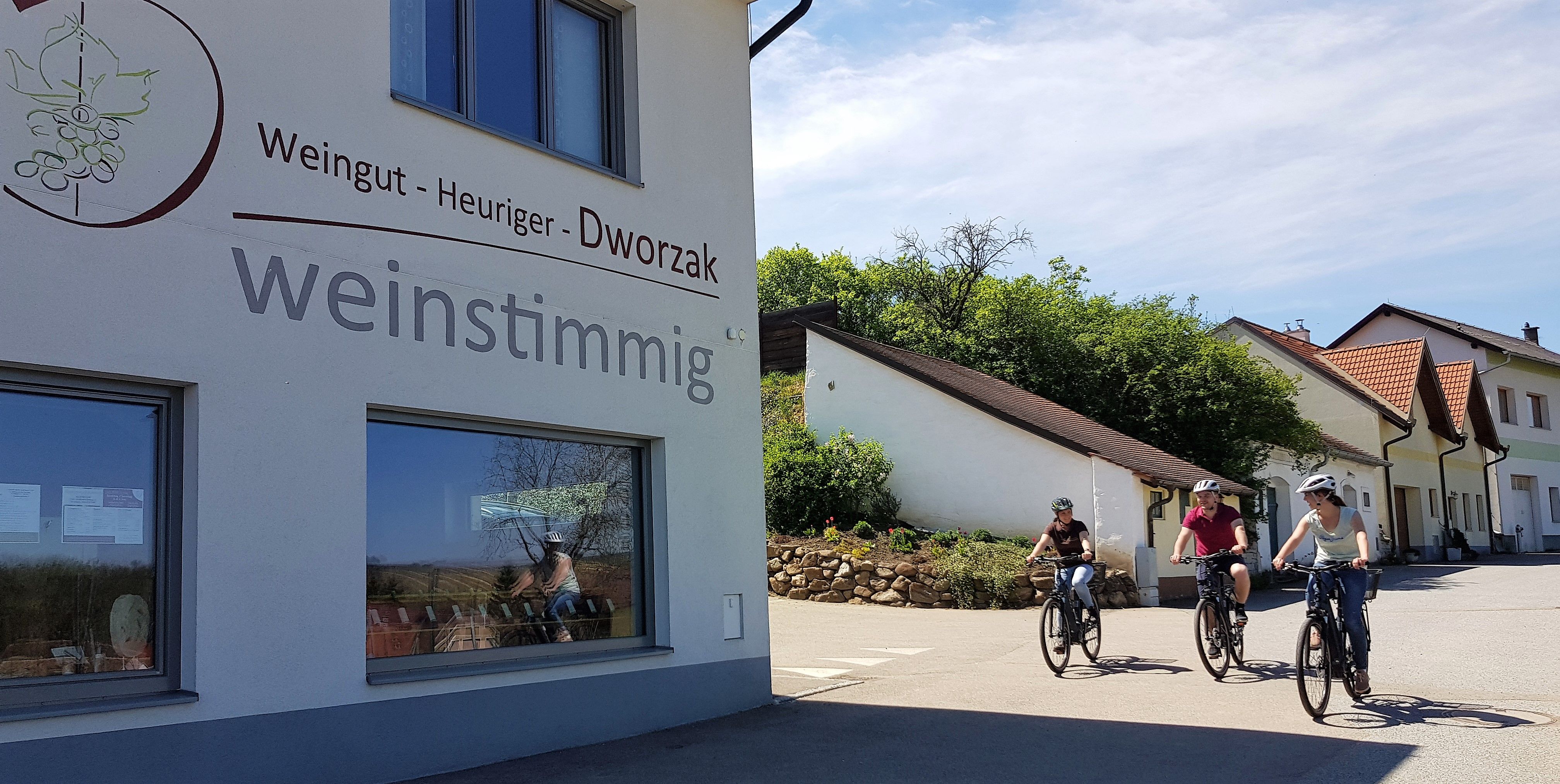 Tři lidé na kolech projíždějí kolem budovy s nápisem "Weingut - heuriger - Dworzak weinstimmig".