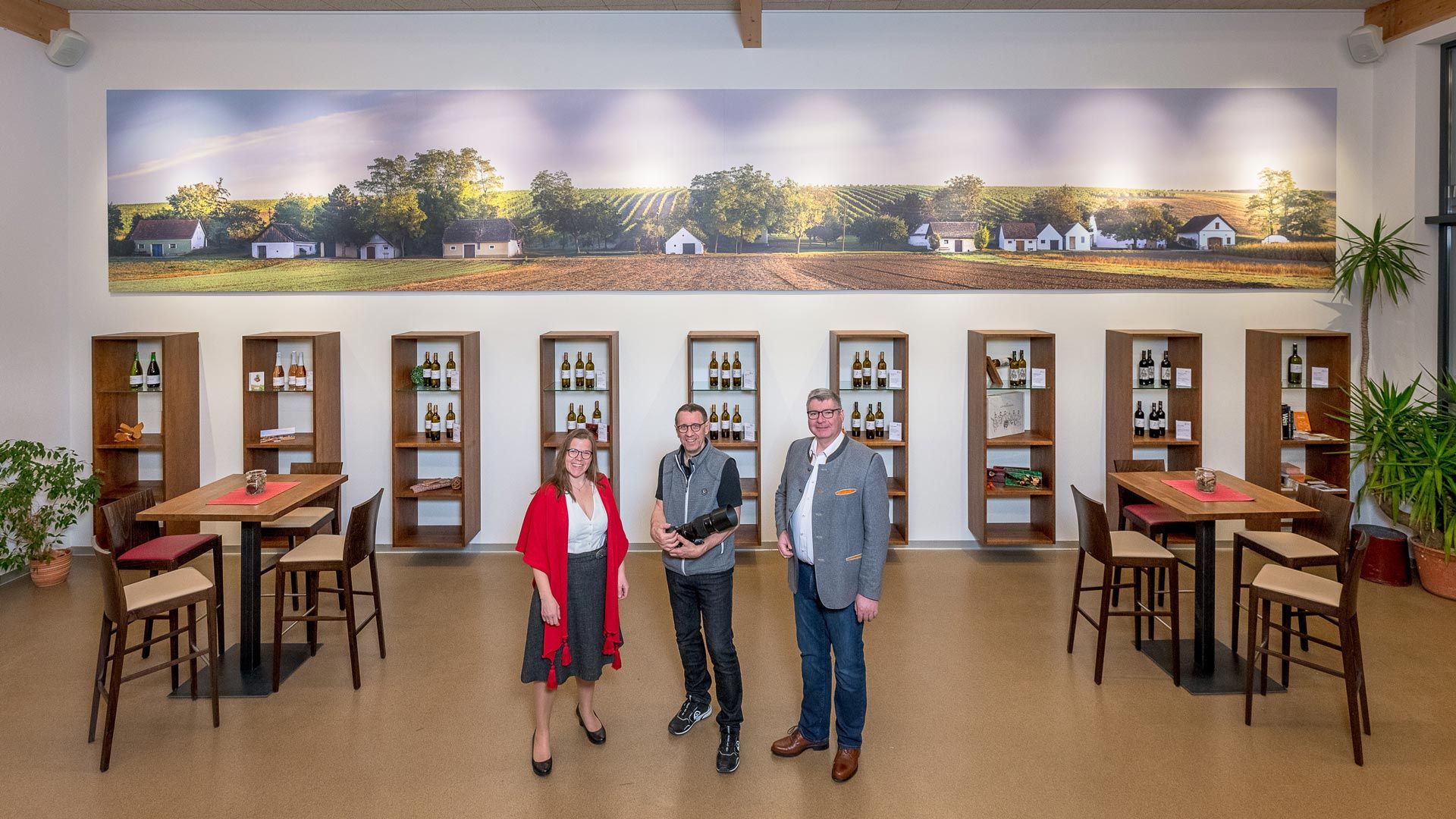 Tři lidé stojí ve vinárně před regály s lahvemi vína a velkou nástěnnou malbou vinic.