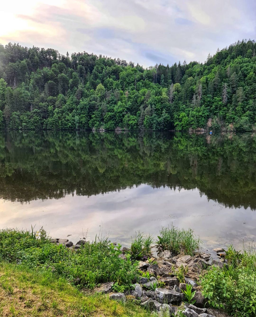 Klidné jezero se zalesněnými kopci v pozadí, které se odrážejí ve vodě.