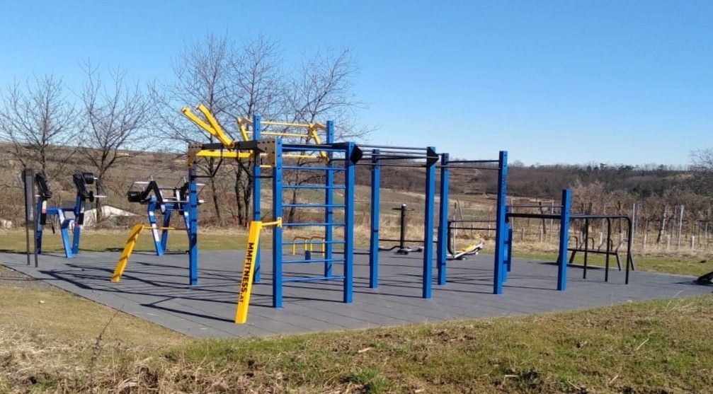 Venkovní kalistenický park s modrými a žlutými prolézačkami, obklopený venkovskou krajinou.