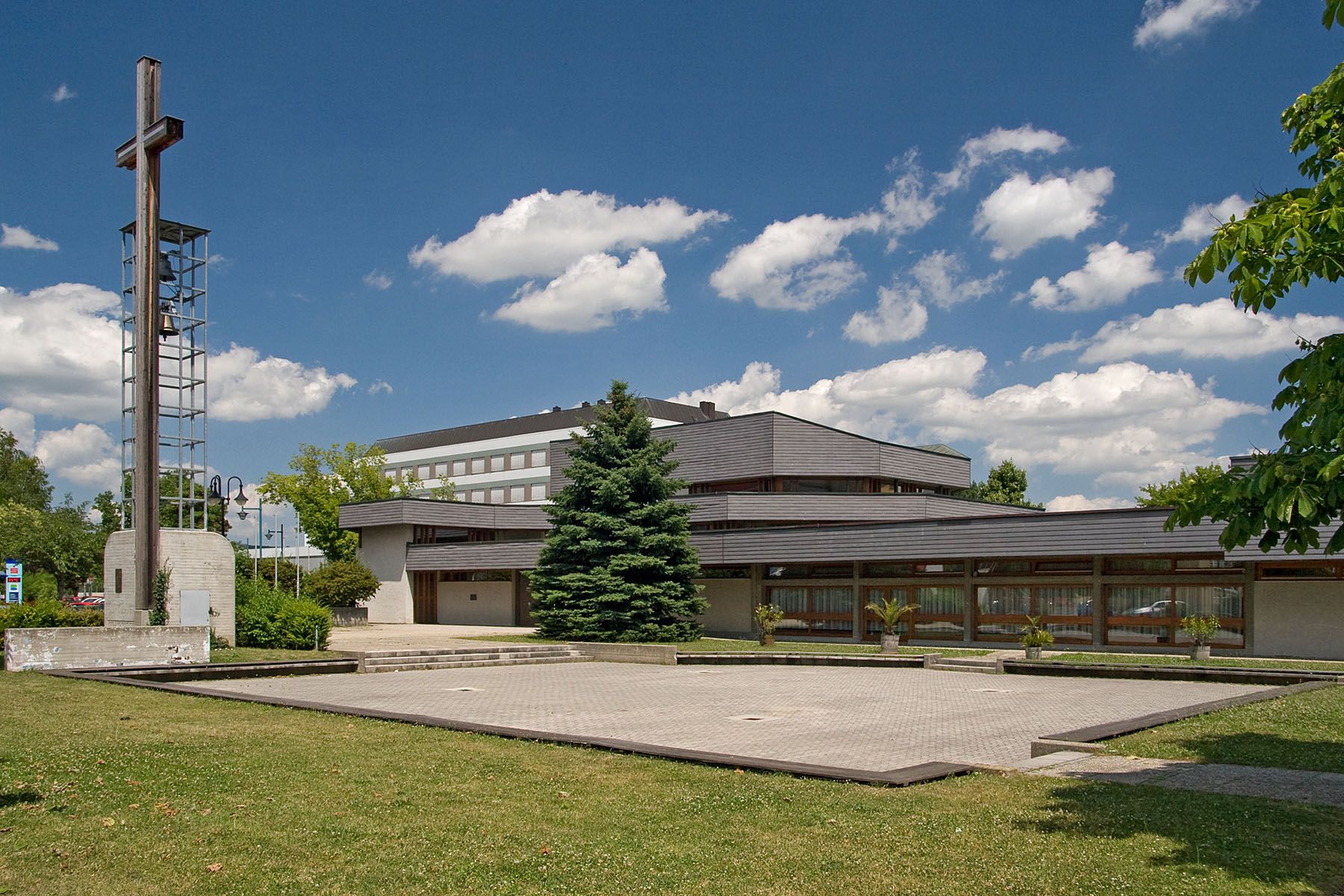 Moderní kostel se zvonicí a křížem, obklopený zelení a stromy, pod modrou oblohou s mraky.