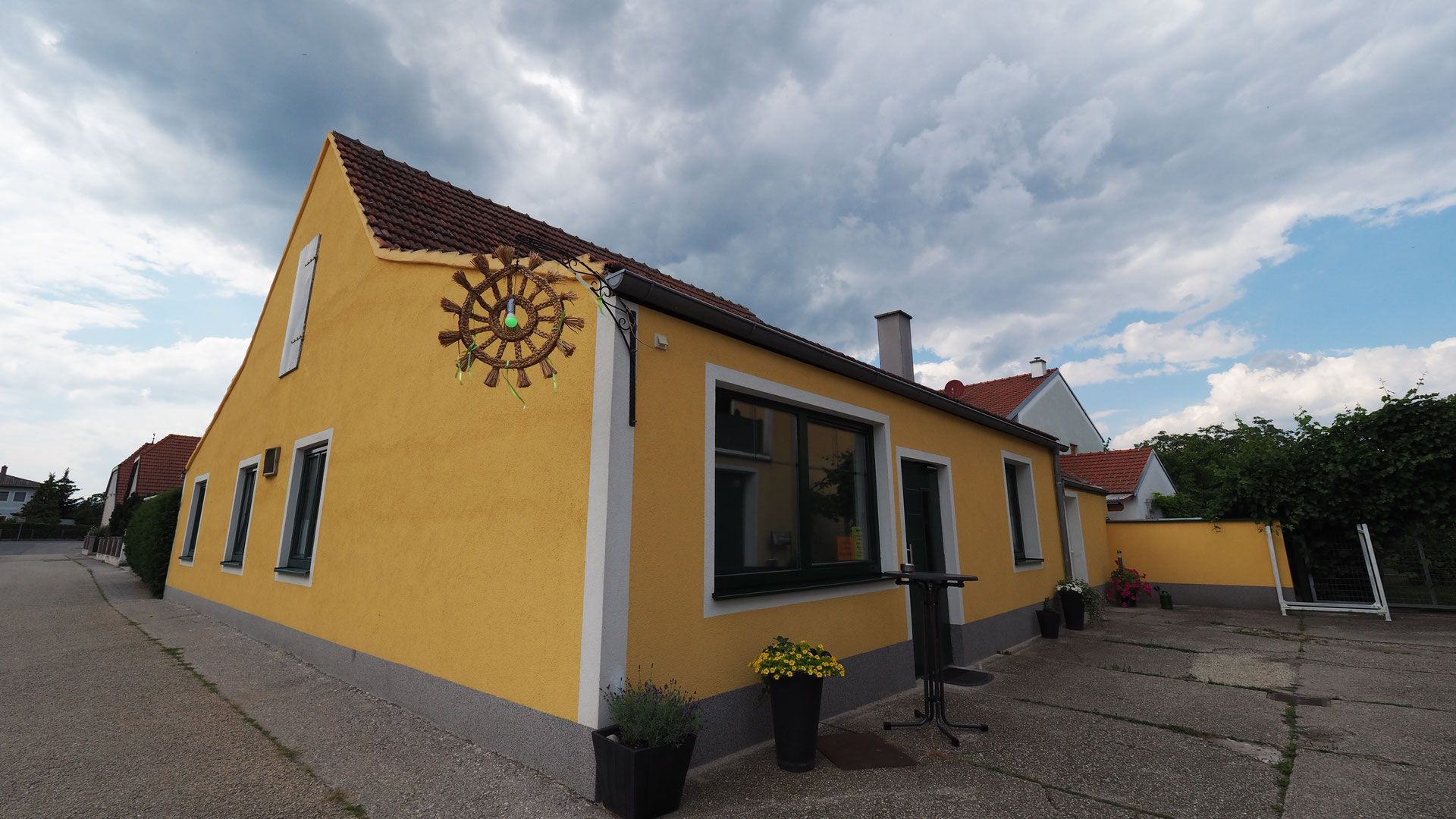 Žlutá budova s červenými střešními taškami a ozdobným kolem na zdi, obklopená rostlinami a zamračenou oblohou.